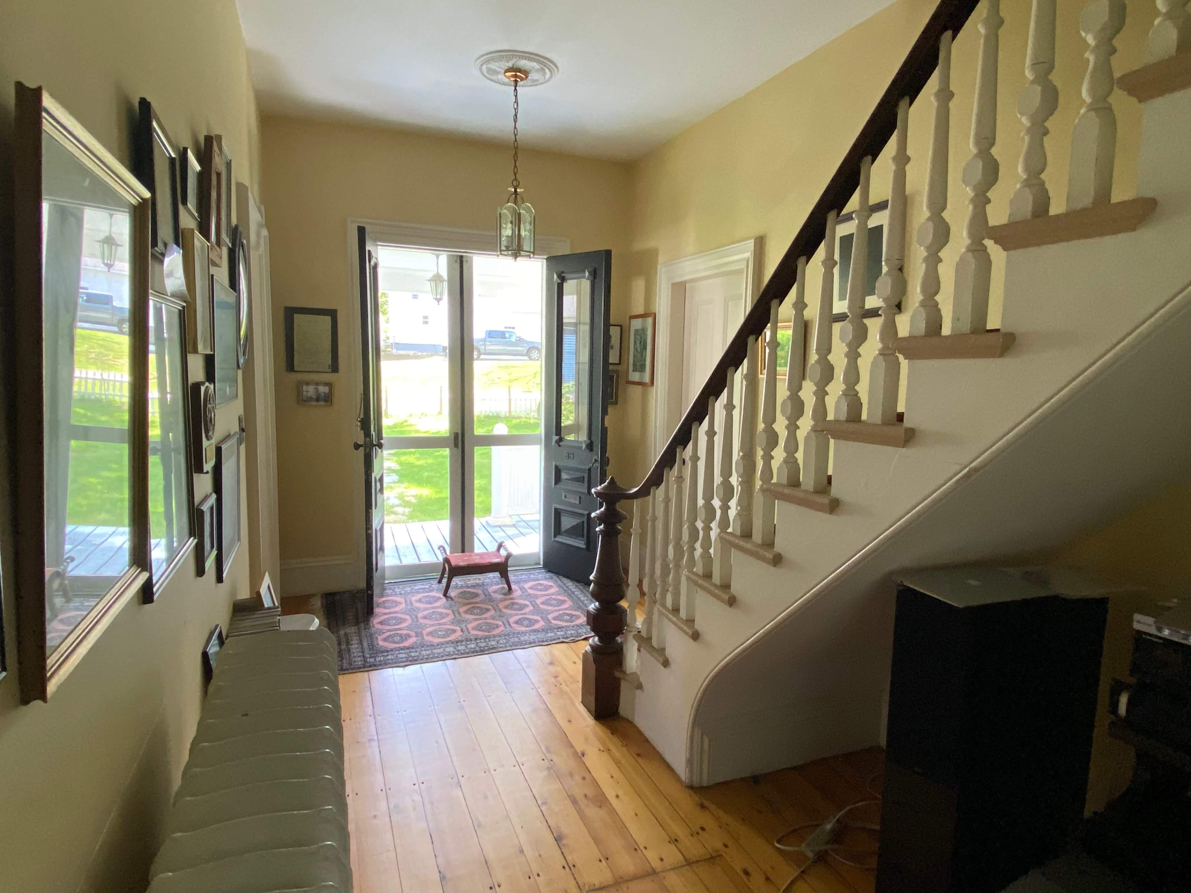 A hallway with a staircase on the right, an open front door leading to the outside, and framed pictures on the walls.