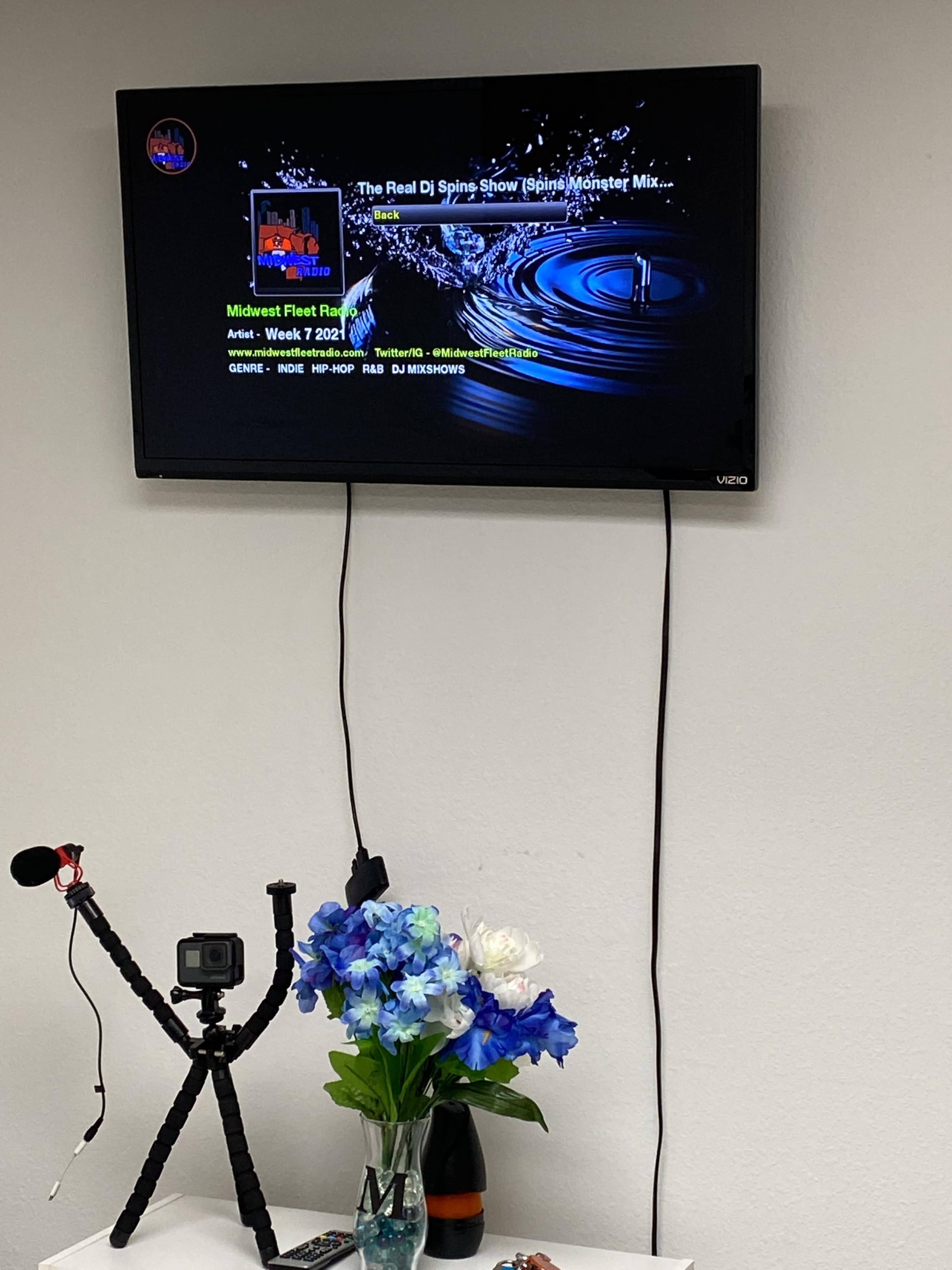 A television displaying promotional artwork for "The Real DJ Spins Show" is mounted on a wall above a table with a camera tripod and a vase of blue flowers.