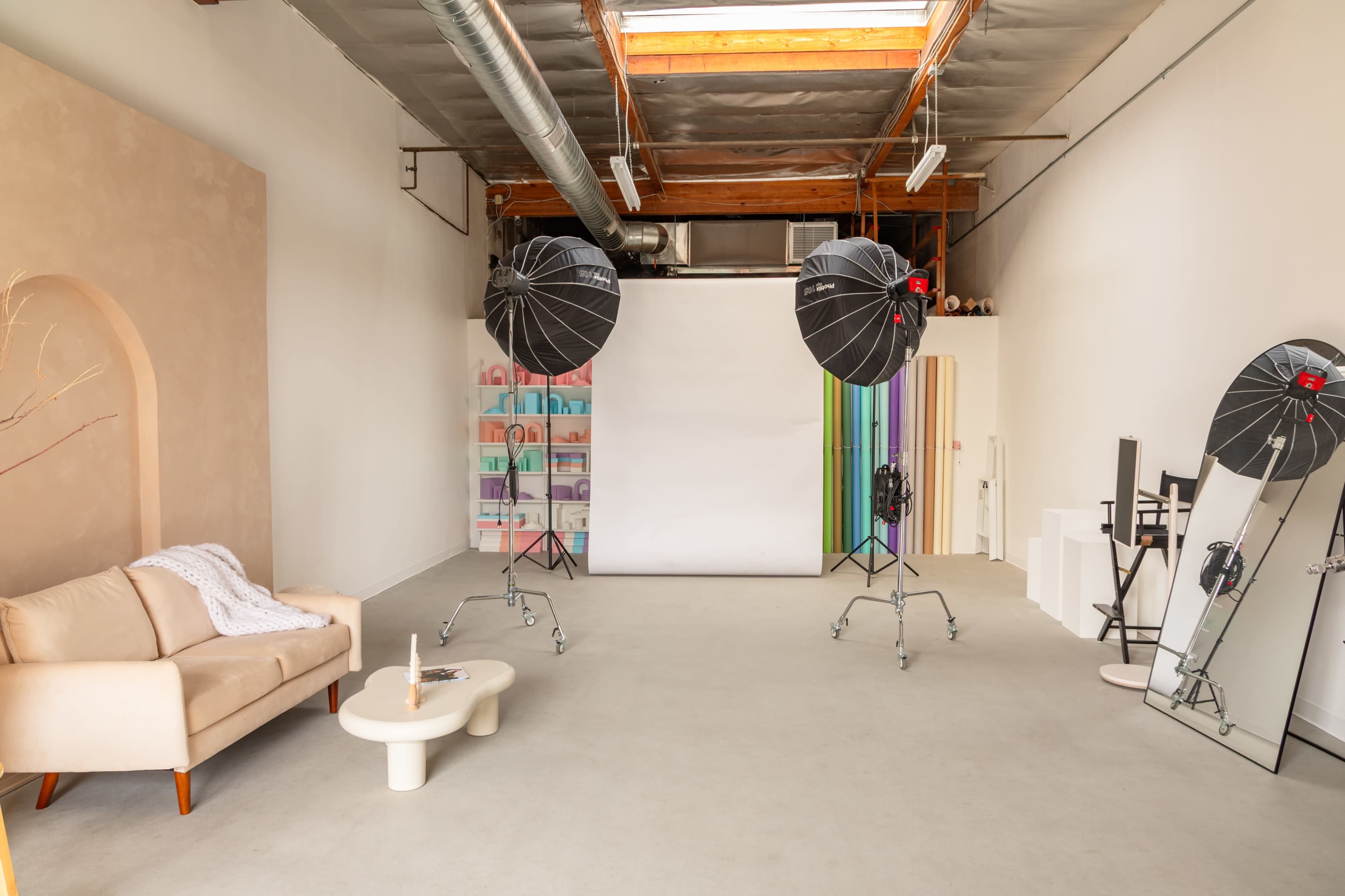 The image shows a spacious photography studio with a cream-colored sofa, large lighting fixtures on stands, a backdrop, and colorful shelves in the background.