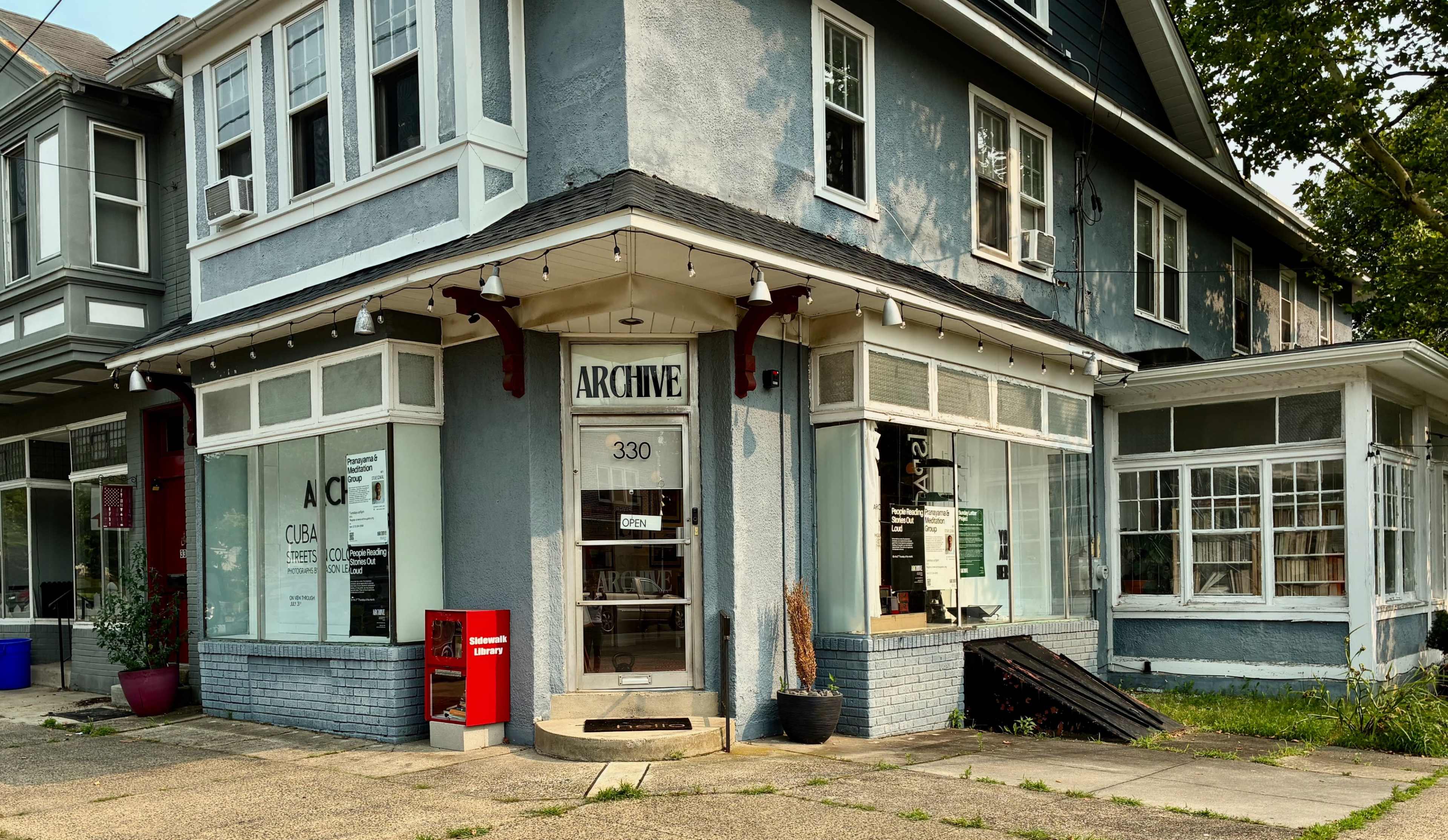The image shows a corner storefront labeled "ARCHIVE" with large windows and a red newspaper box outside, situated on a gray multi-level building.