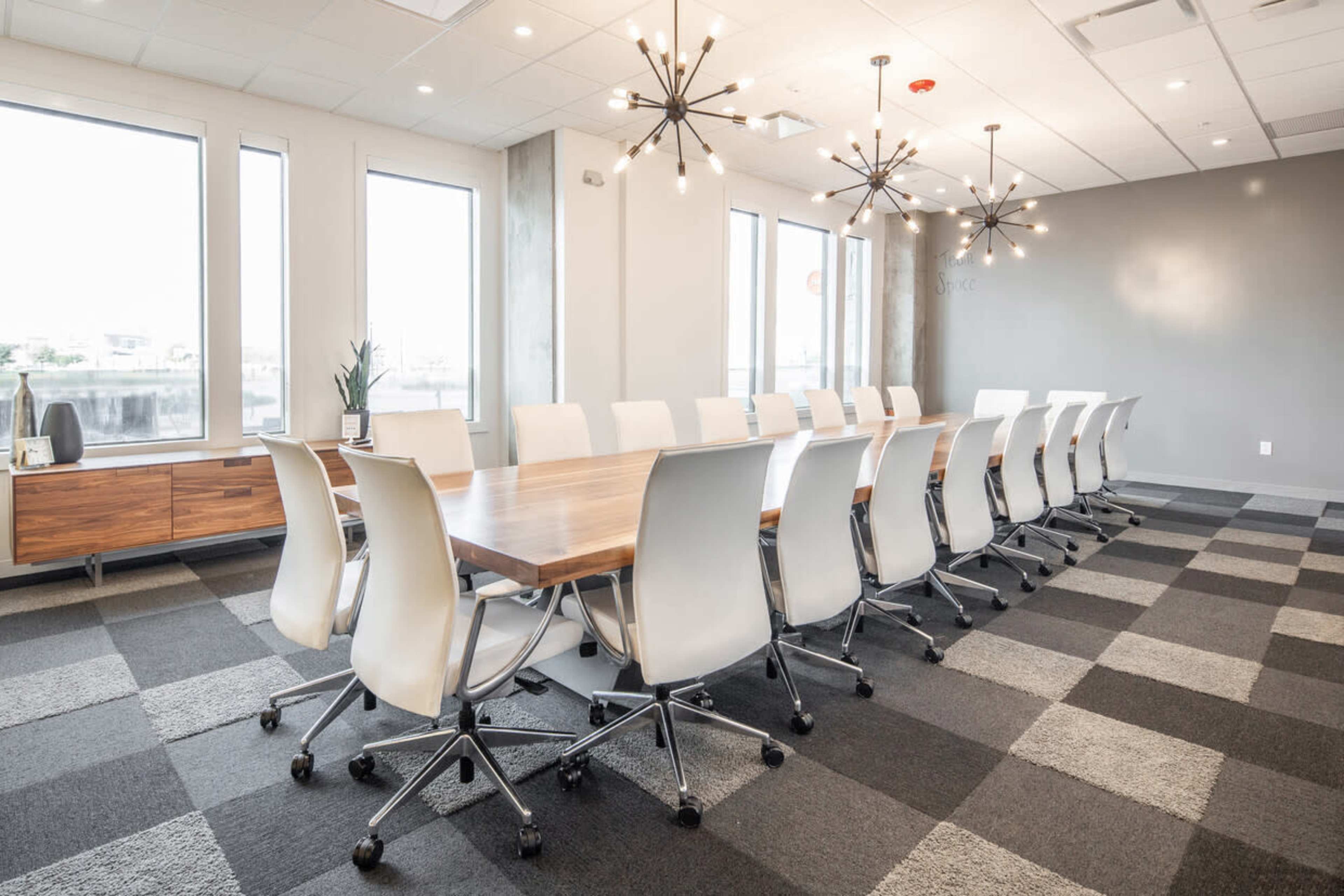 A modern conference room features a long wooden table surrounded by white swivel chairs, with large windows and contemporary light fixtures overhead.