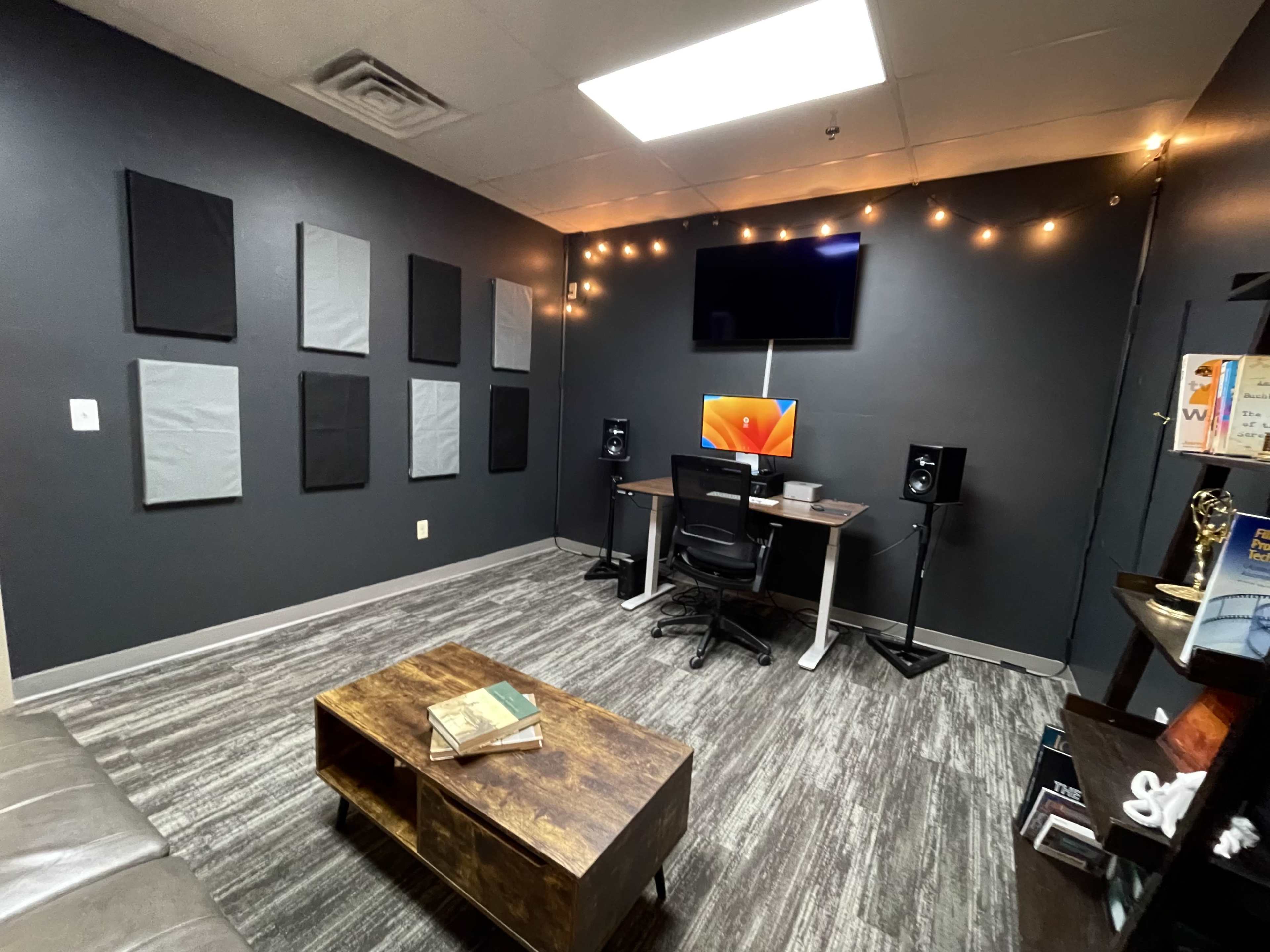 A modern office space with gray walls, a wooden coffee table, a desk setup with a computer and speakers, and acoustic panels on the walls.