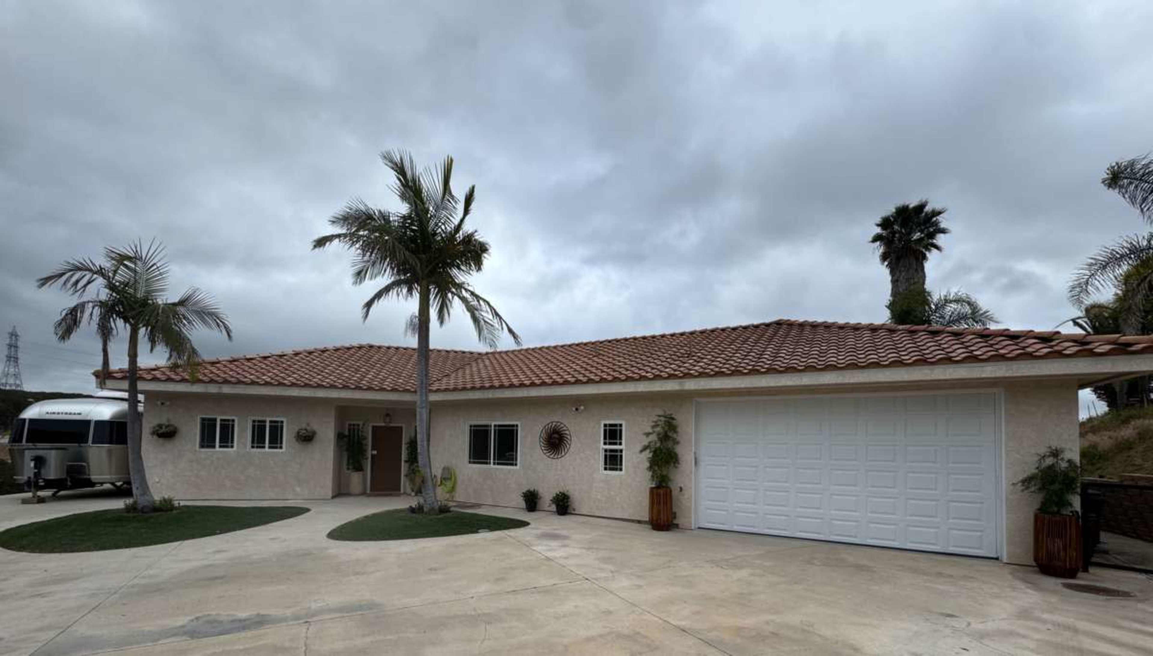 A single-story house with a tiled roof, palm trees, and a large garage door sits under a cloudy sky.