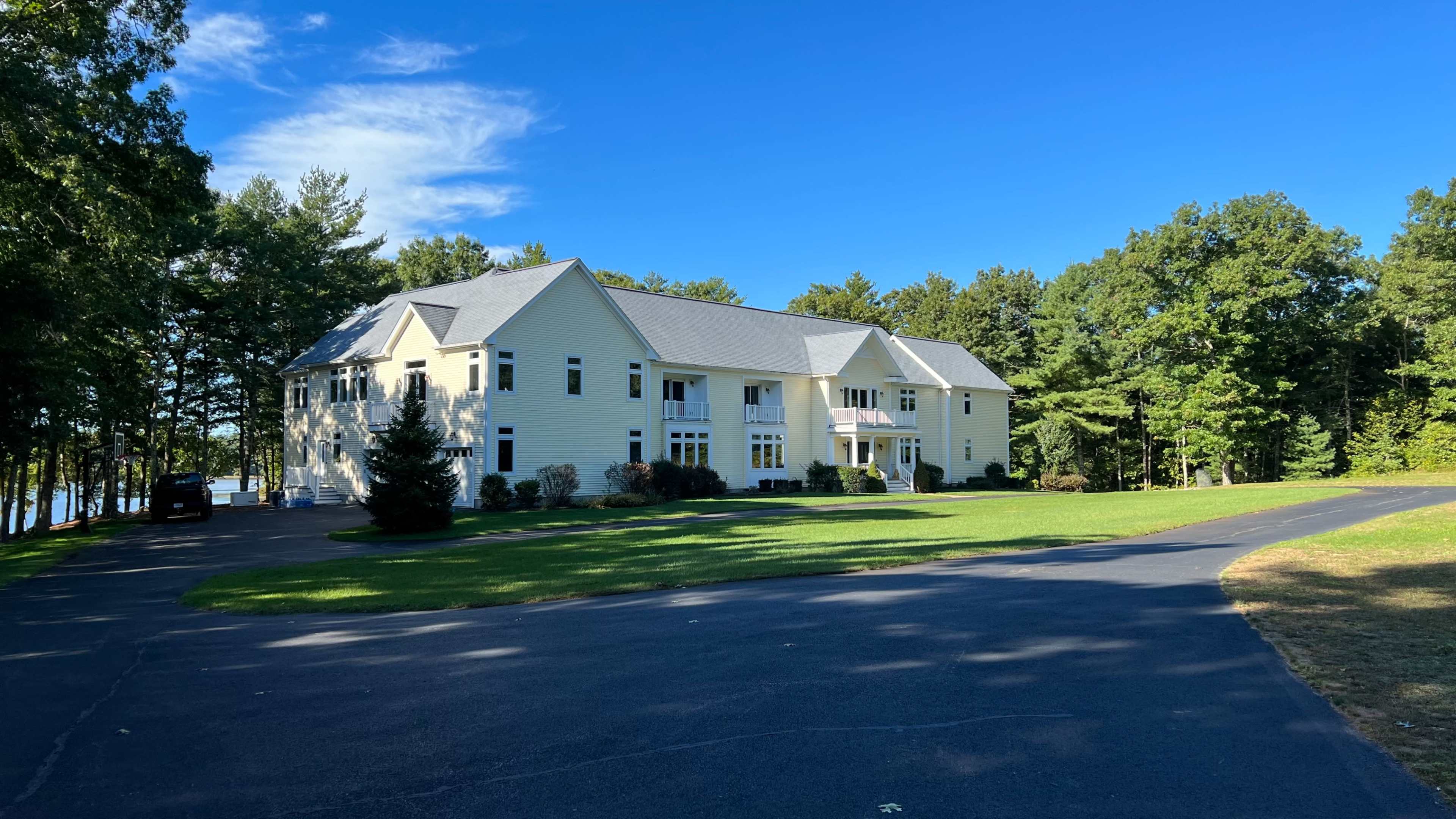 A large yellow house with multiple balconies sits on a manicured lawn beside a tree line and a paved driveway.