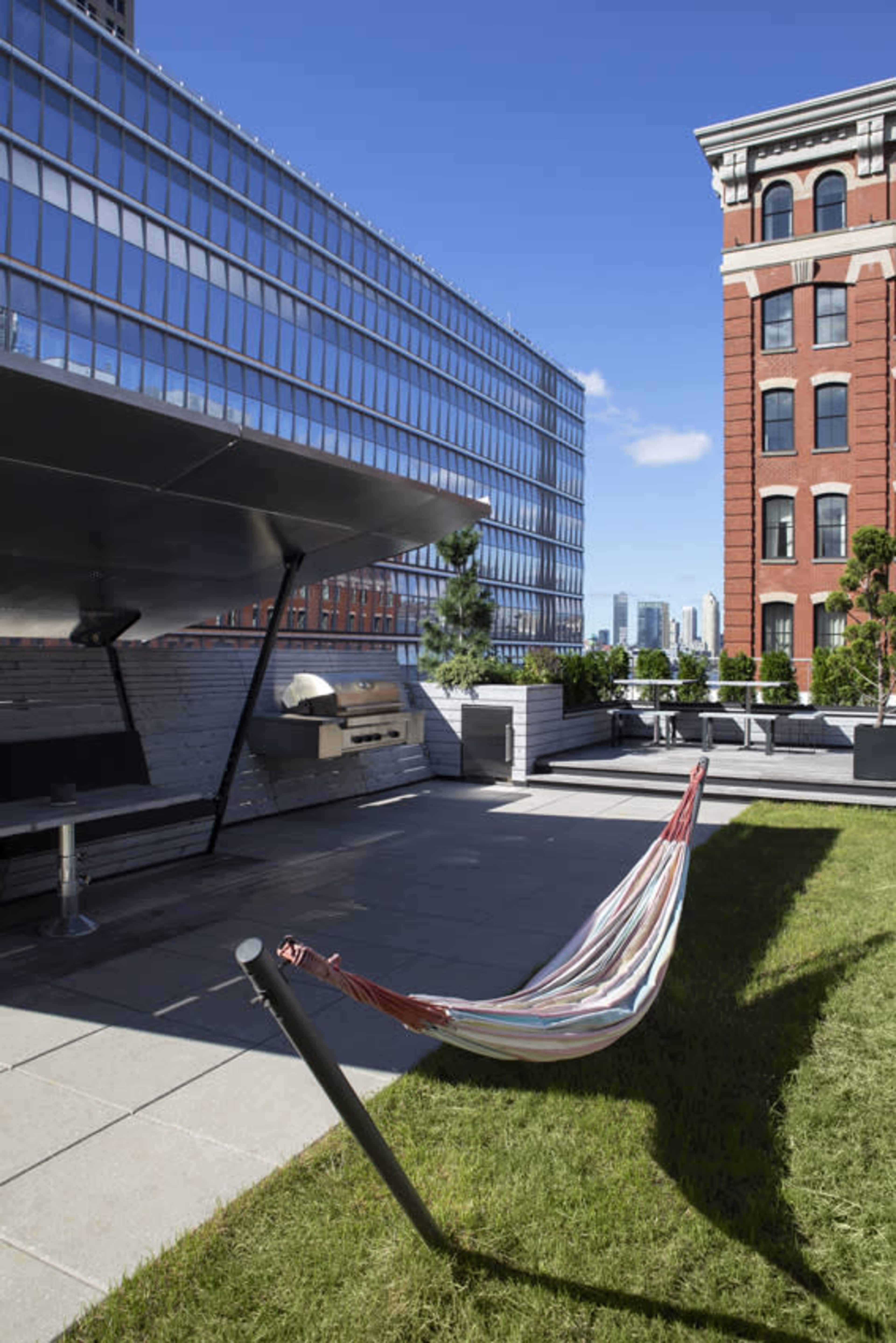 A striped hammock is suspended between two supports on a rooftop terrace, surrounded by modern buildings and greenery.