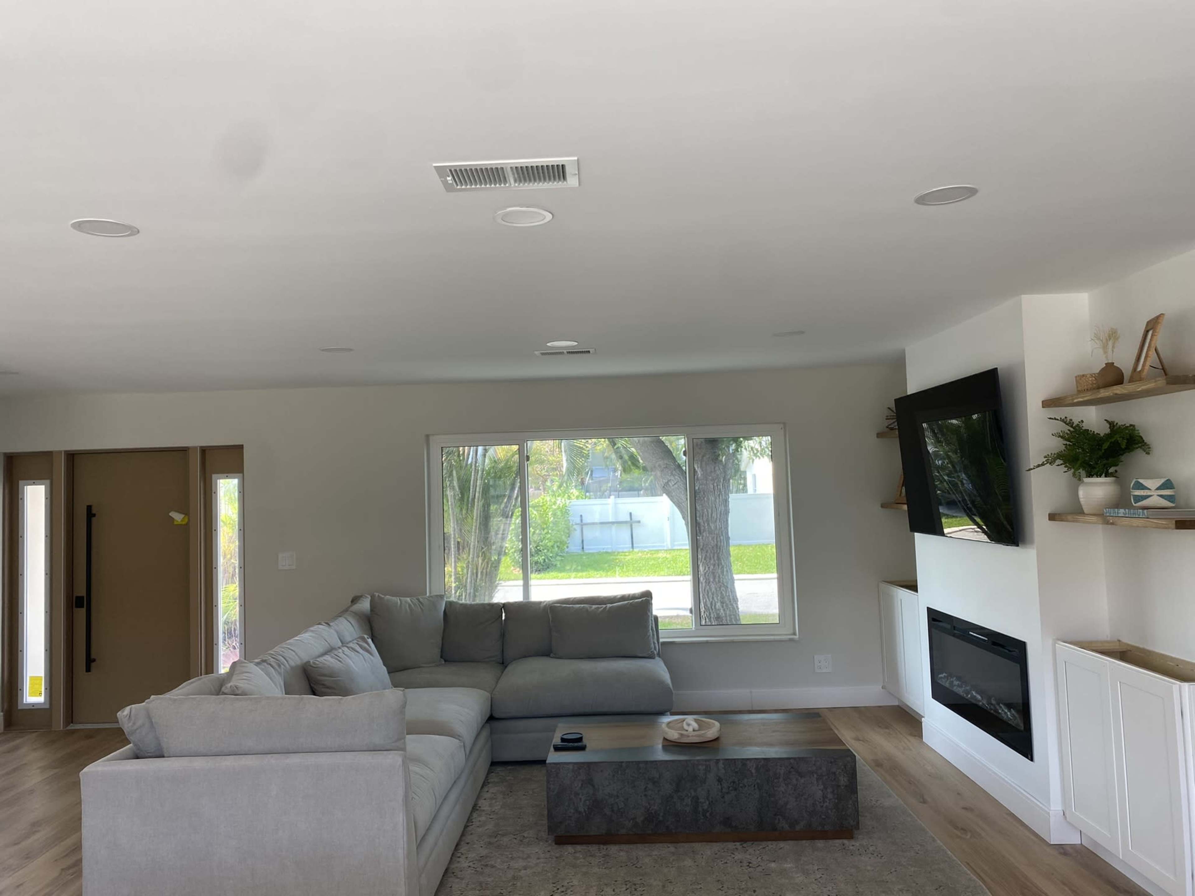 The image shows a modern living room with a large sectional sofa, a coffee table, a television mounted on the wall, and a fireplace beneath it, along with a window providing natural light.