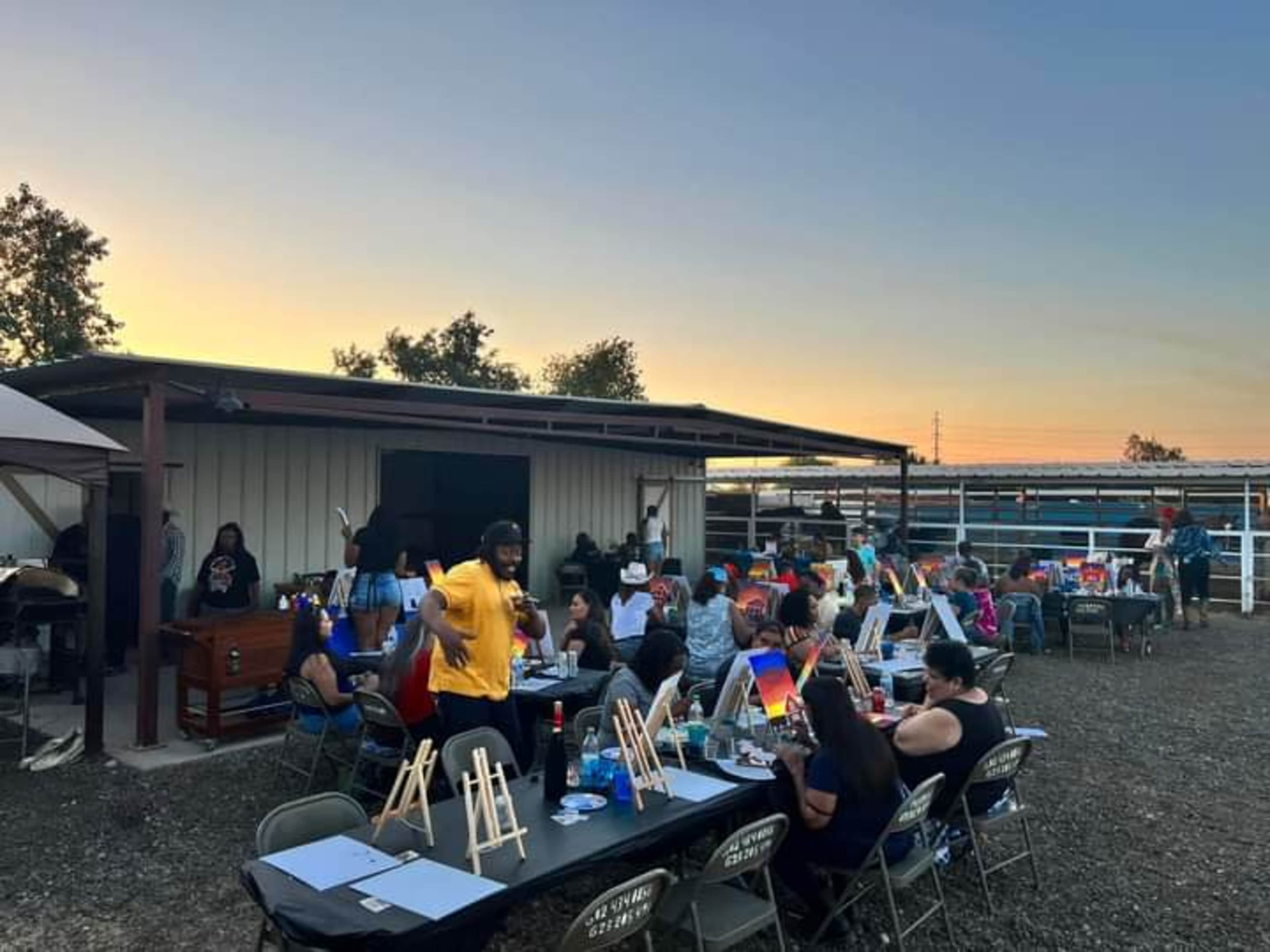 A group of people participates in a painting event outdoors, surrounded by easels and art supplies as the sun sets in the background.