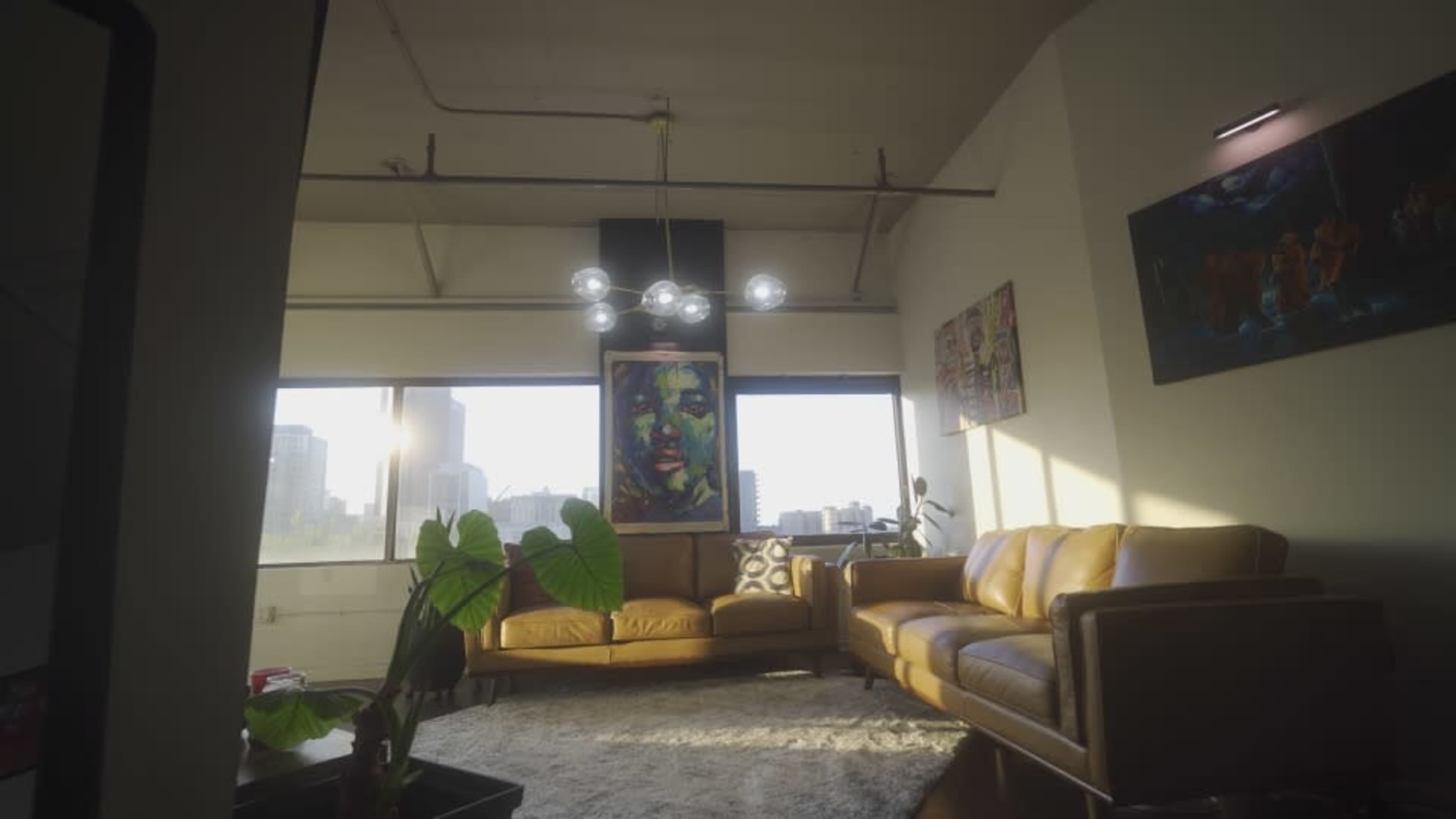 A modern living room features a leather couch, vibrant artwork, large windows with city views, and a decorative plant.