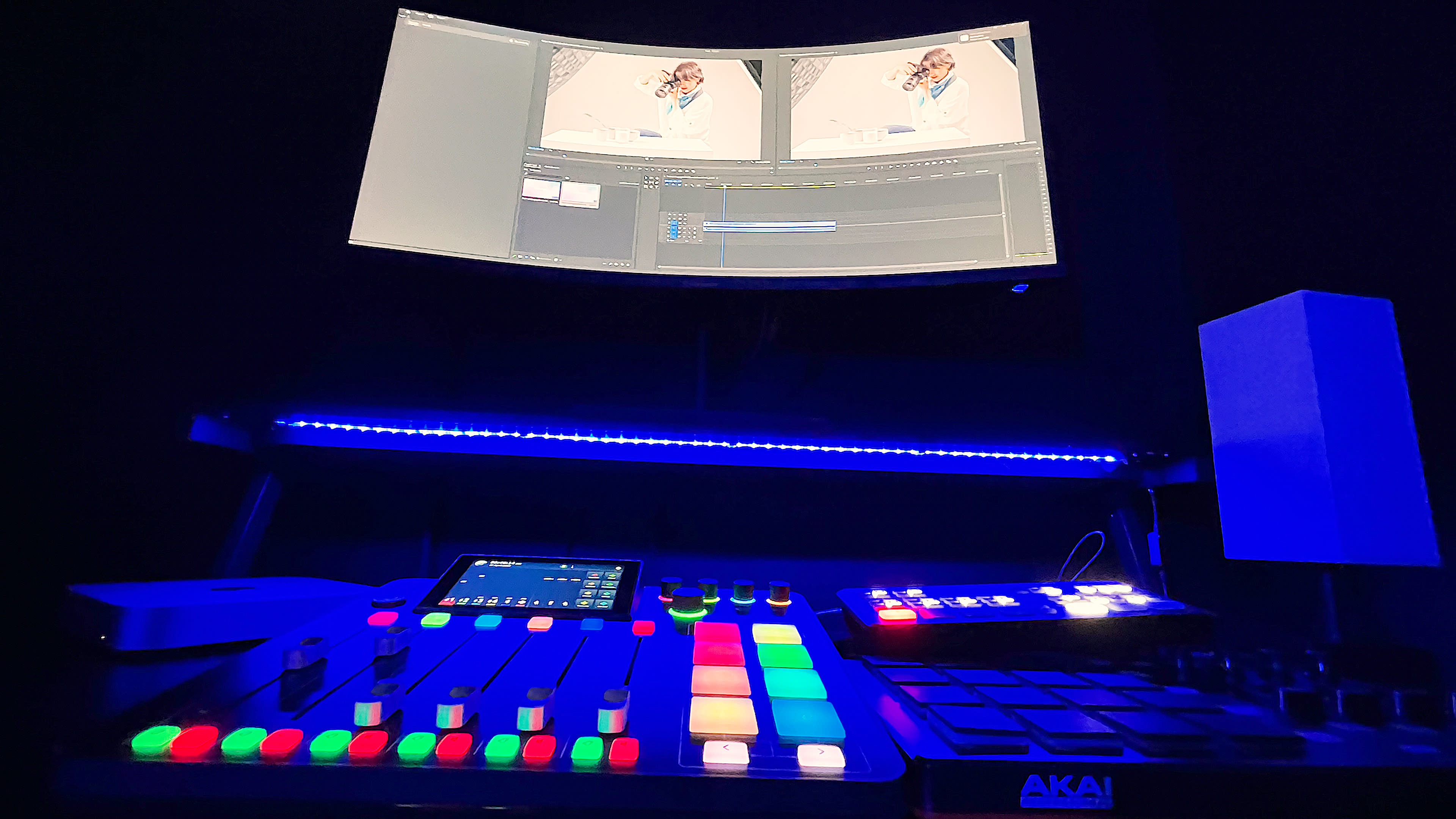 The image shows a music production setup with a mixer, a touchscreen controller, and a large curved monitor displaying a video editing interface.