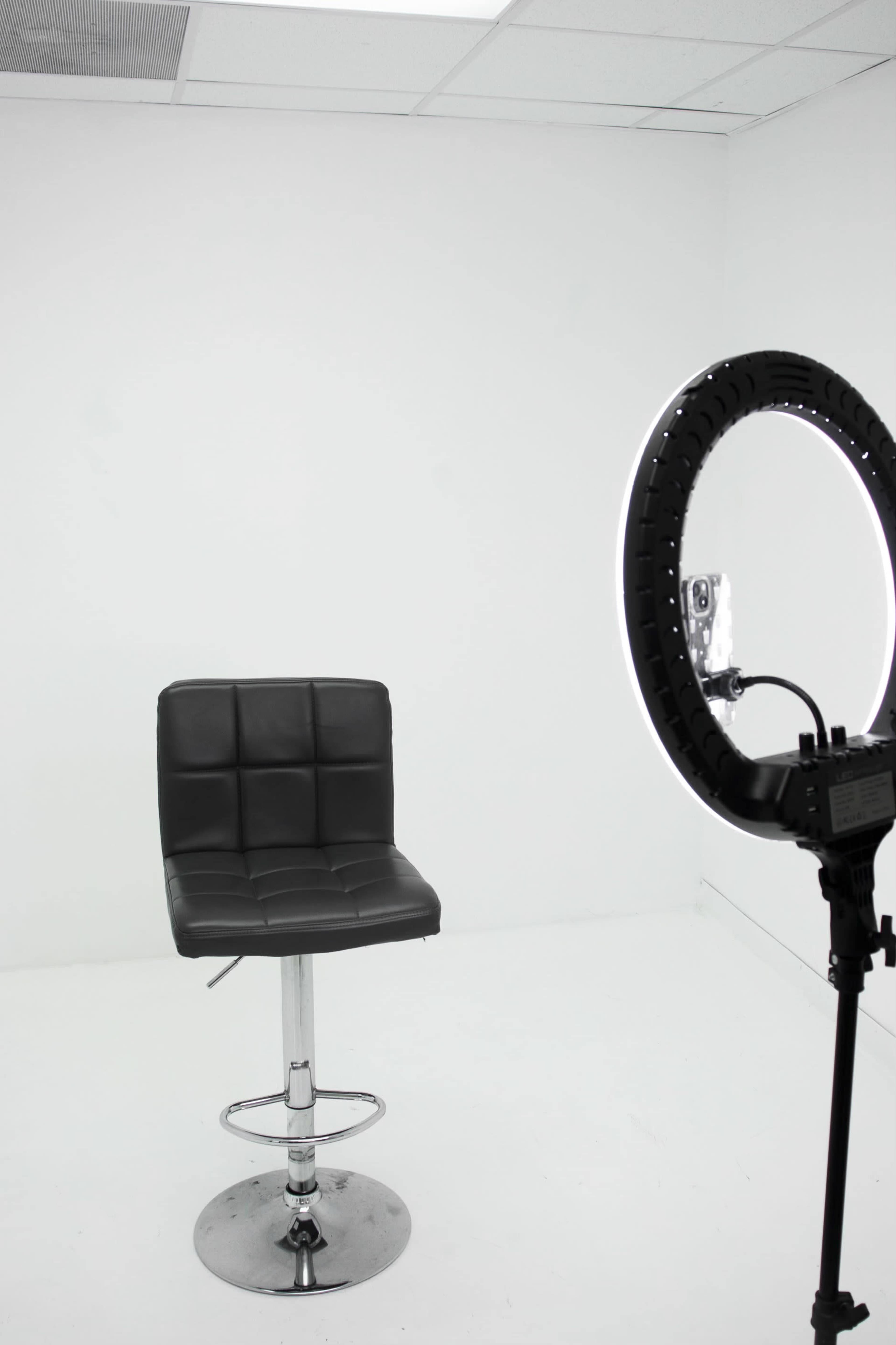 A black leather bar stool is positioned in a minimalist white studio next to a large ring light on a stand.