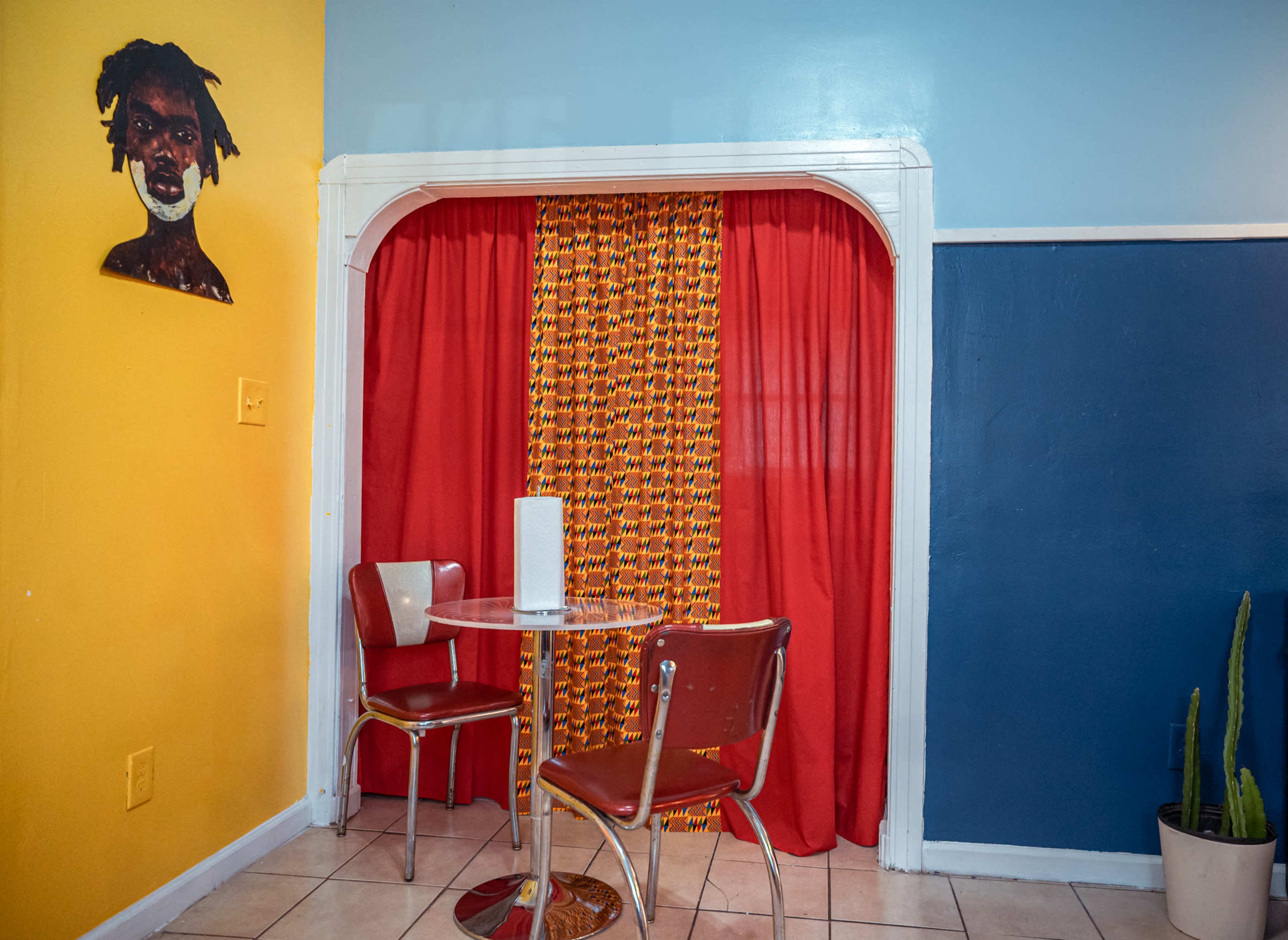 A small seating area features two red chairs and a round table in front of a colorful curtain, with a painting on the yellow wall.