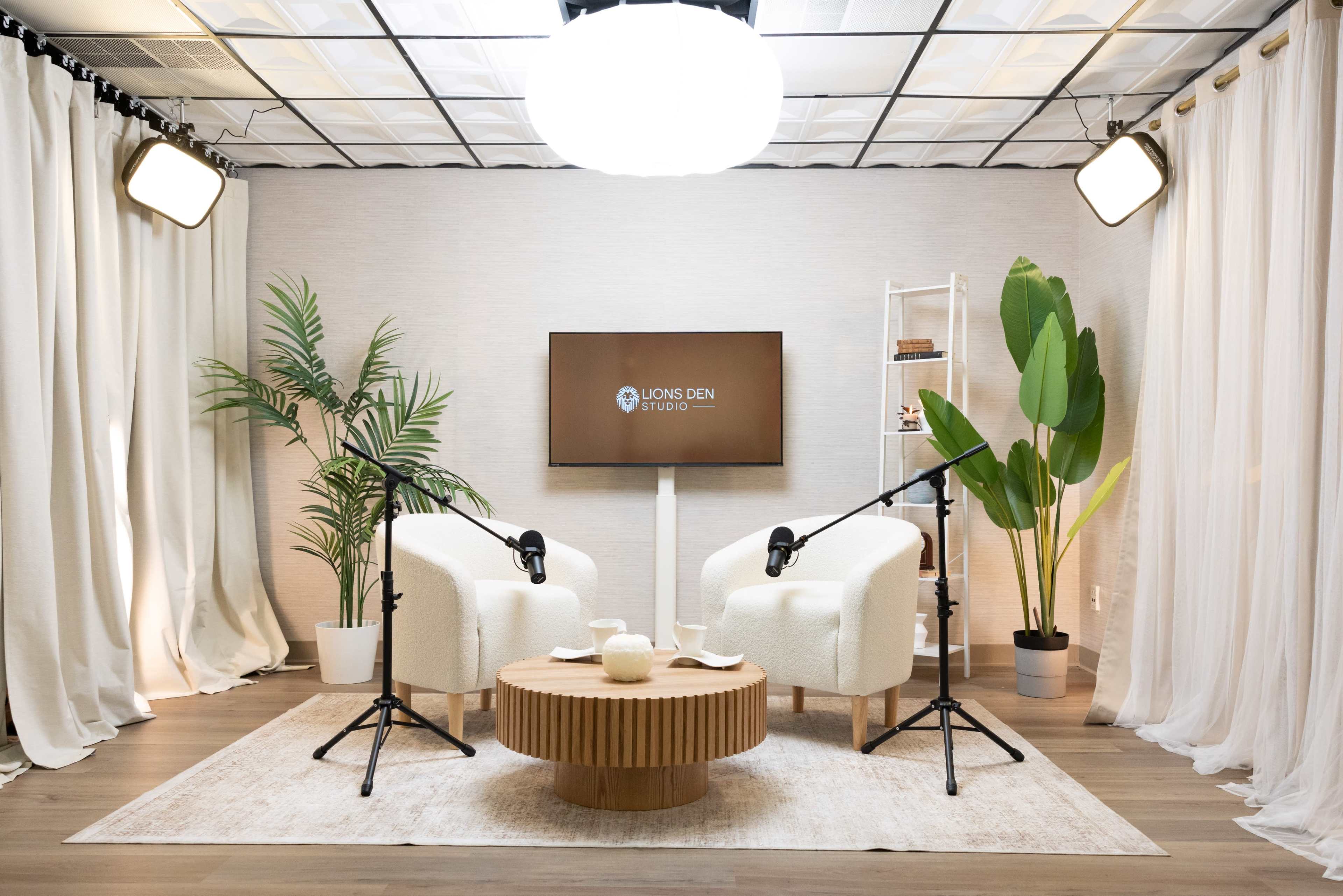 The image shows a modern studio setup with two white armchairs facing a screen, surrounded by plants and lit by overhead lights.