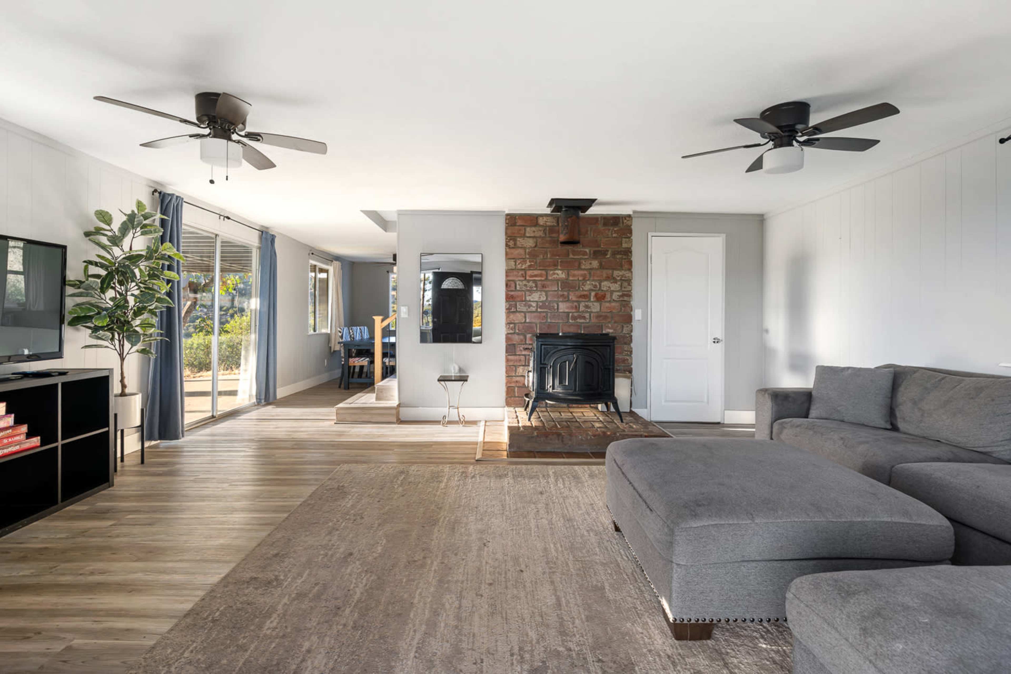 The image shows a spacious living room featuring a gray sectional sofa, a brick fireplace with a wood stove, and two ceiling fans.
