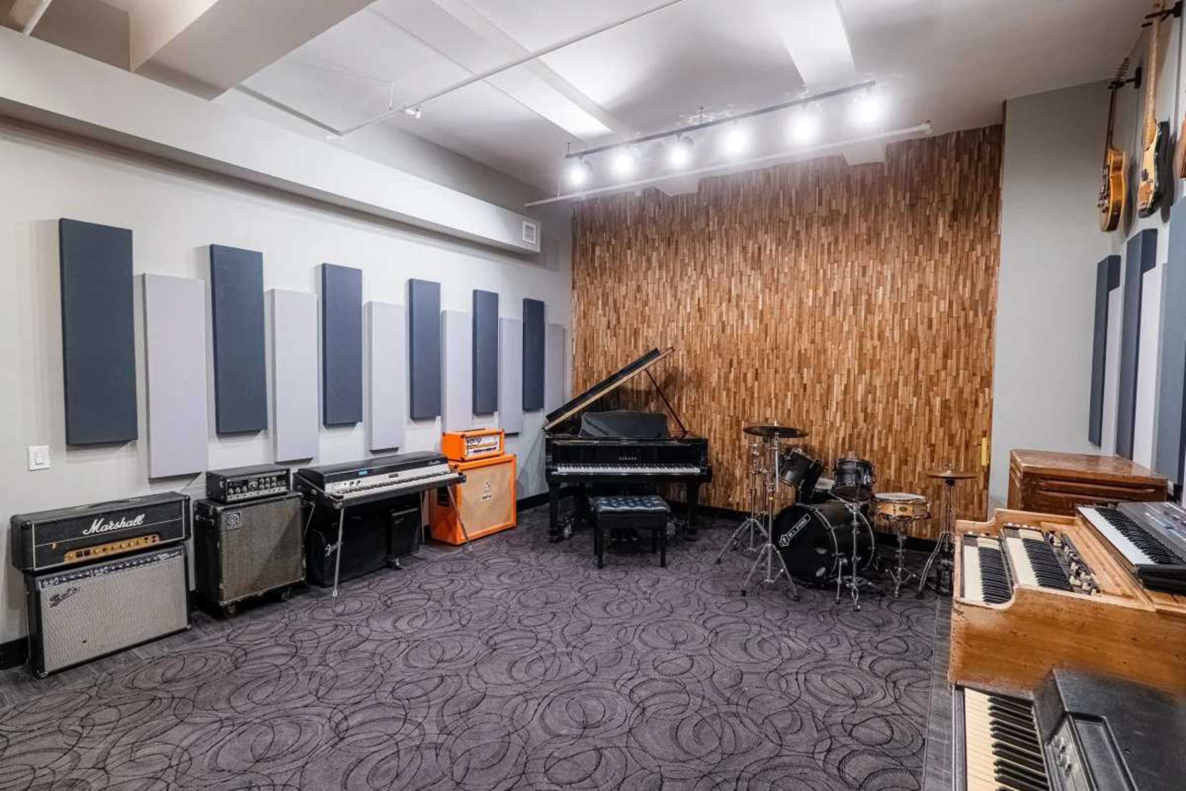 The image shows a music studio equipped with various instruments, including amplifiers, a grand piano, a drum set, and synthesizers, arranged in an acoustically treated room.