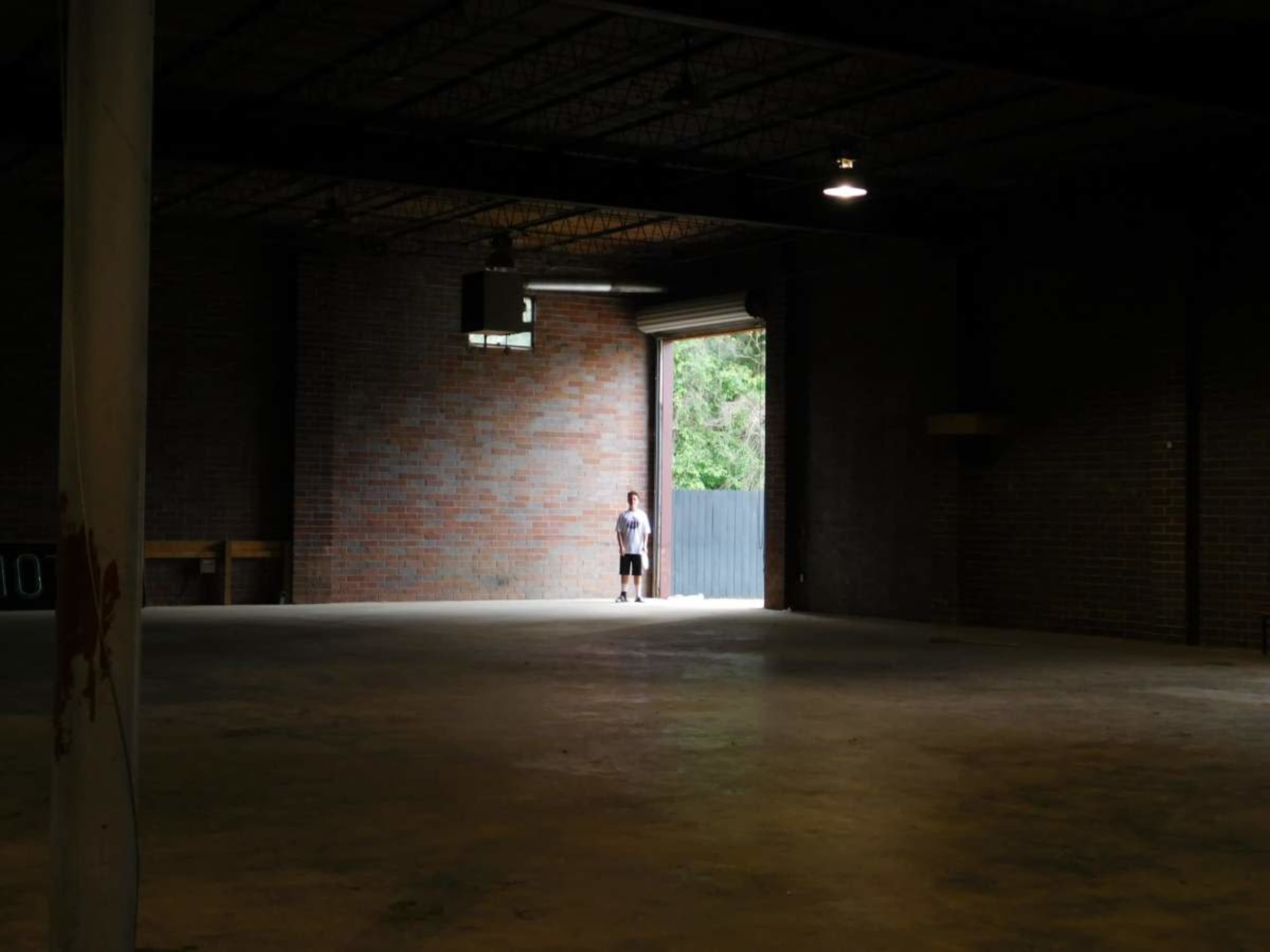 A person stands alone in a large, empty room with brick walls and a doorway leading outside.