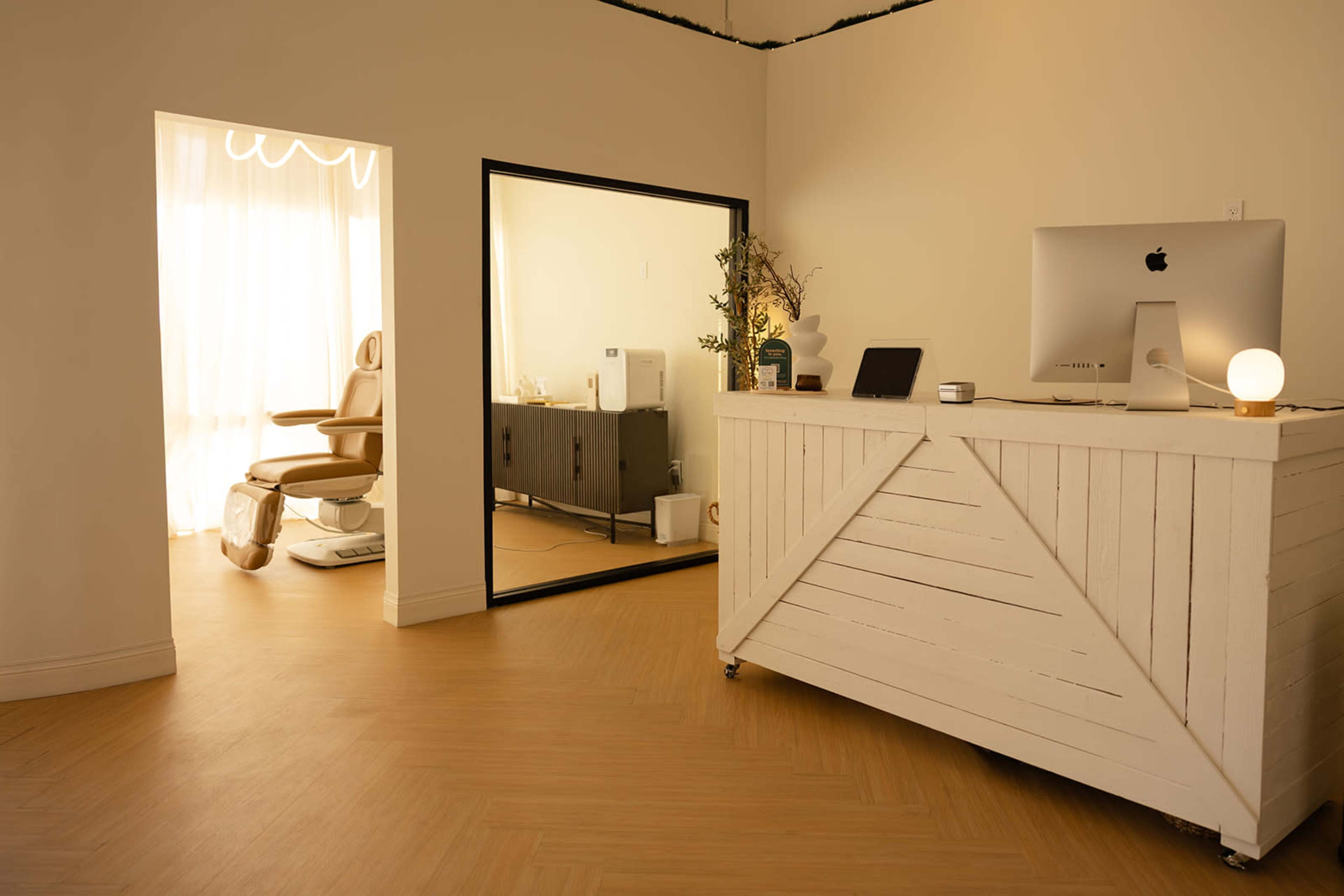 The image shows a minimalist room featuring a reception desk made of white wood, a large mirror reflecting a treatment chair and a dark cabinet, with natural light coming in from a window.
