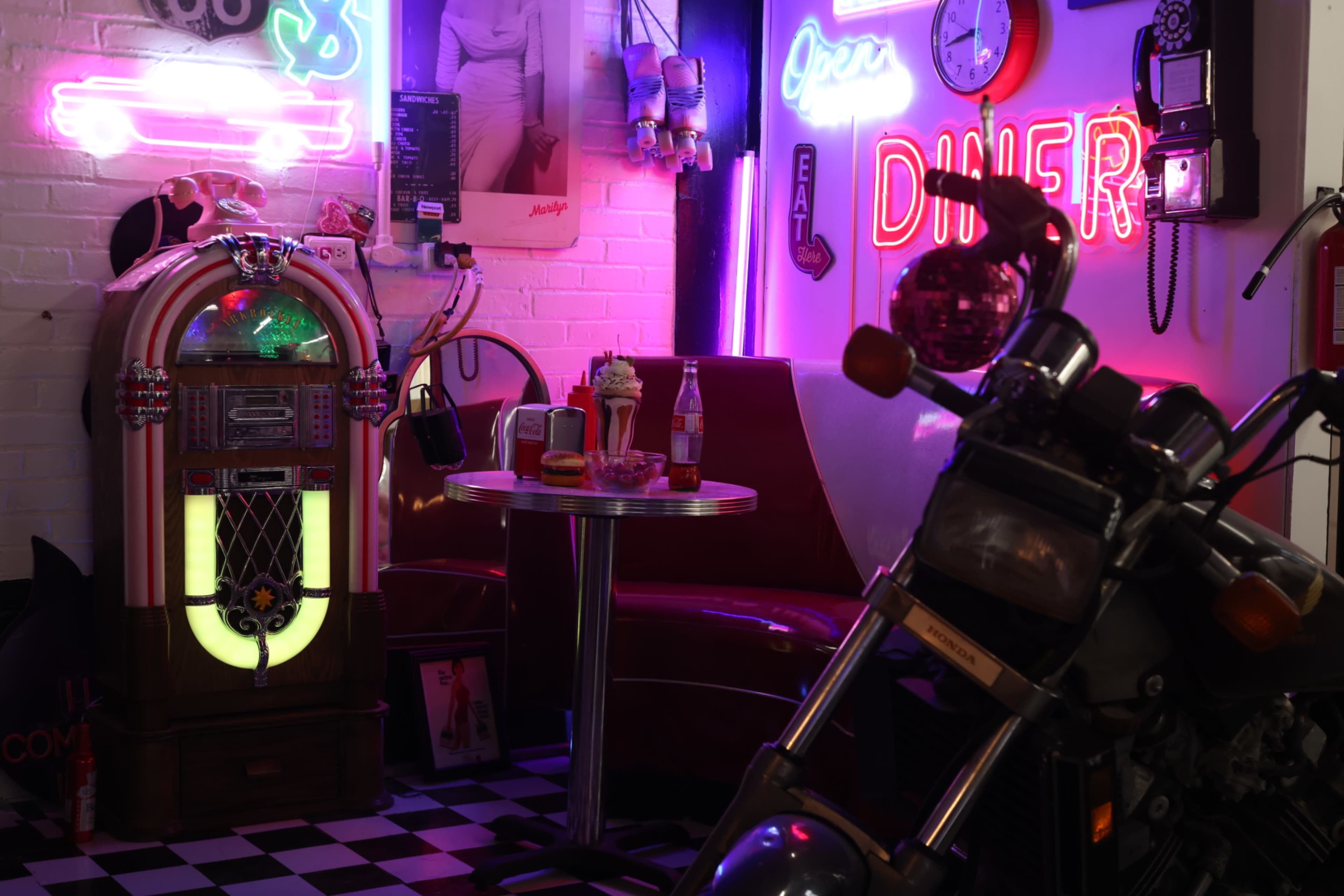 The image depicts a retro diner scene featuring a jukebox, a booth with a table, neon lights, and a motorcycle parked nearby.