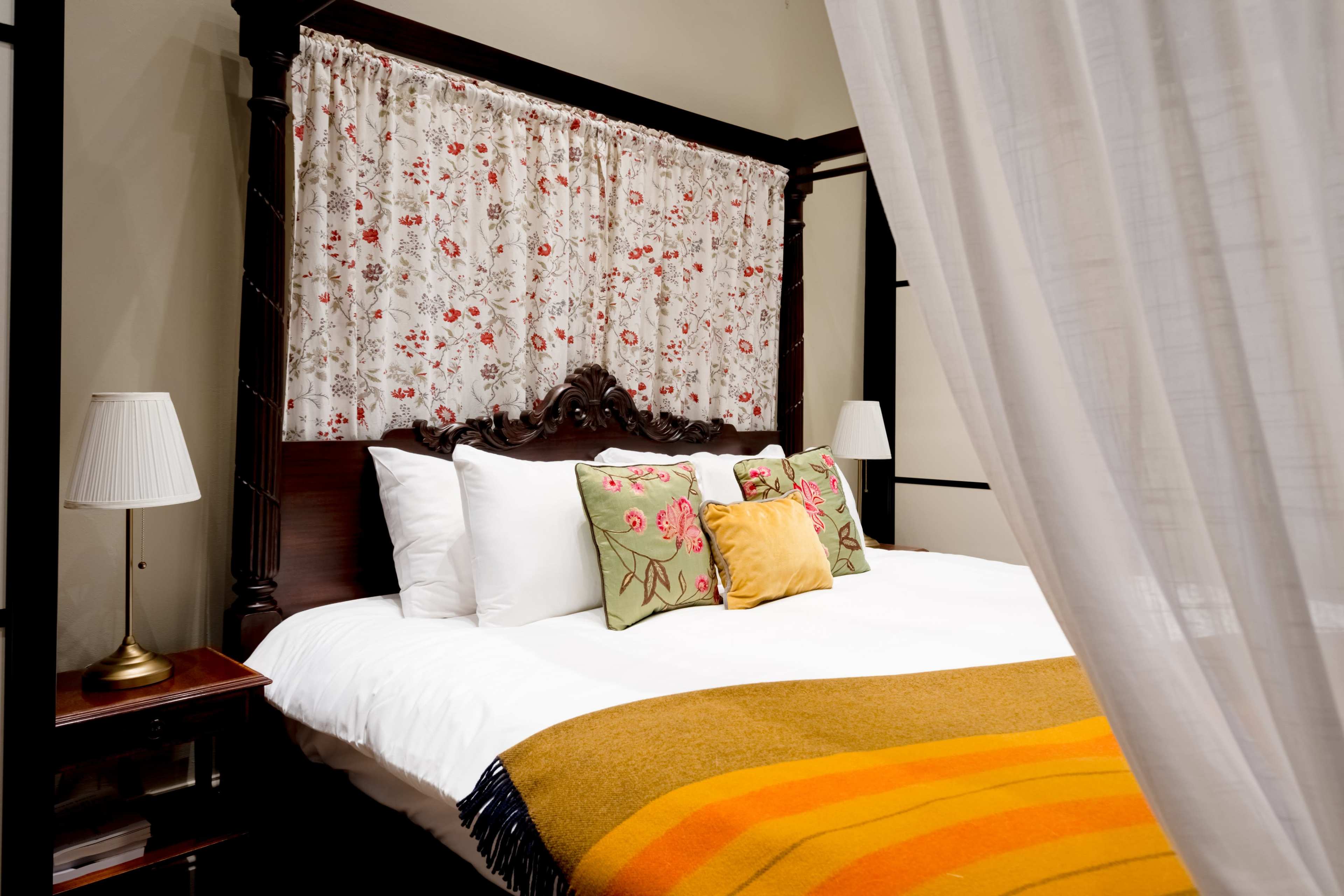 The image shows a canopy bed with a floral curtain, colorful pillows, and a blanket draped over the edge in a softly lit room.