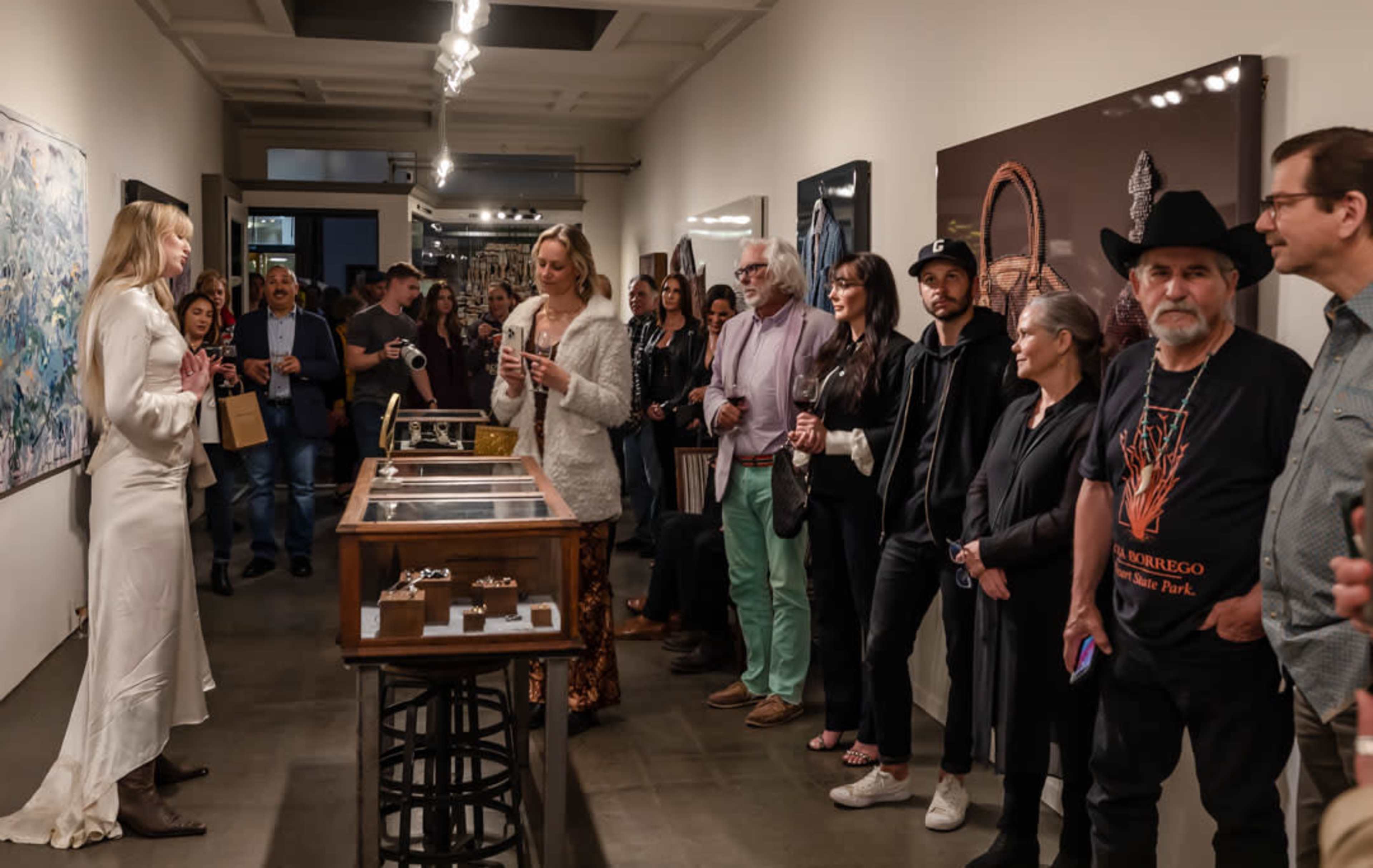 A group of people stands attentively in an art gallery, interacting with a presenter near a display of jewelry.