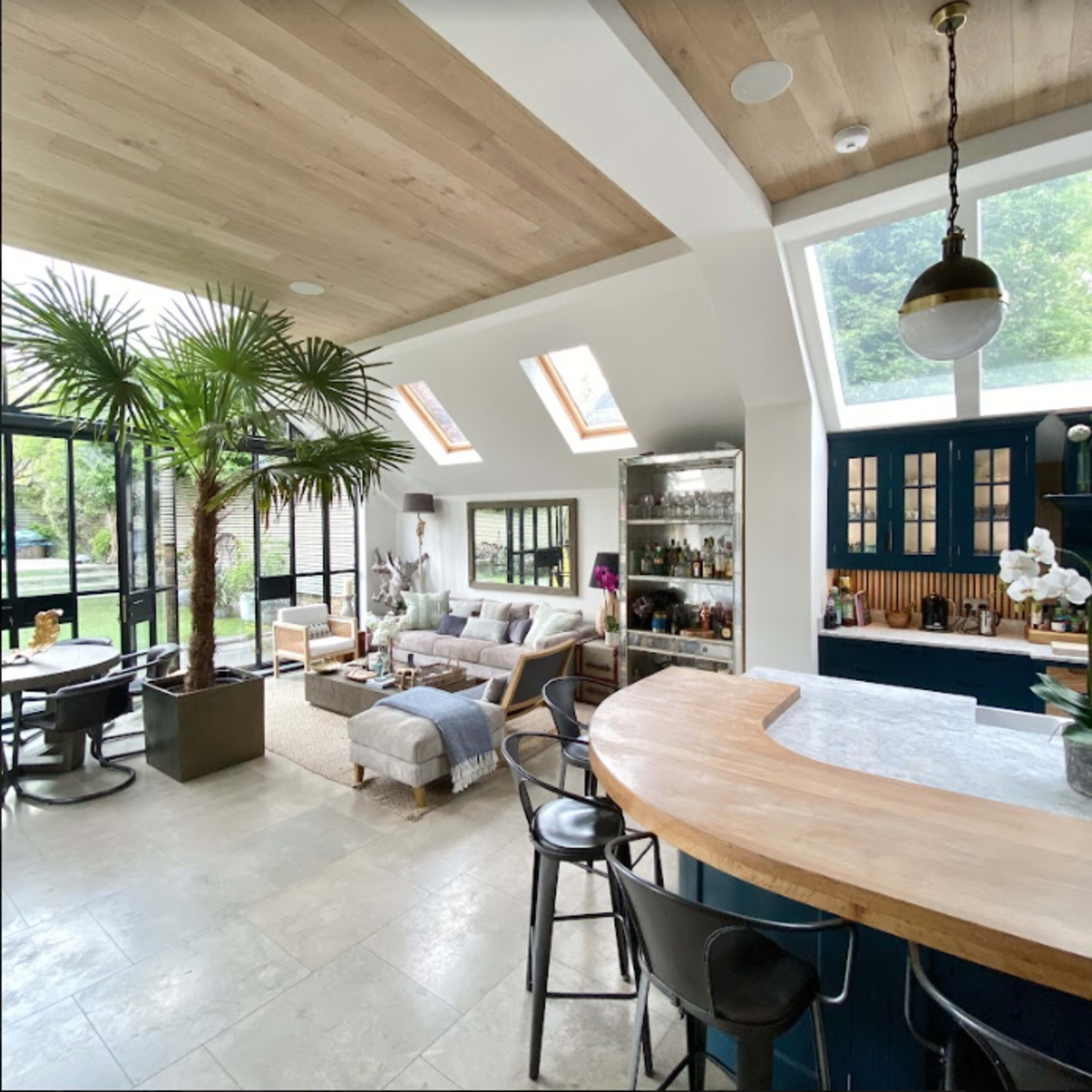 A bright, modern living space featuring a cozy seating area, a bar with stools, large windows, and greenery.