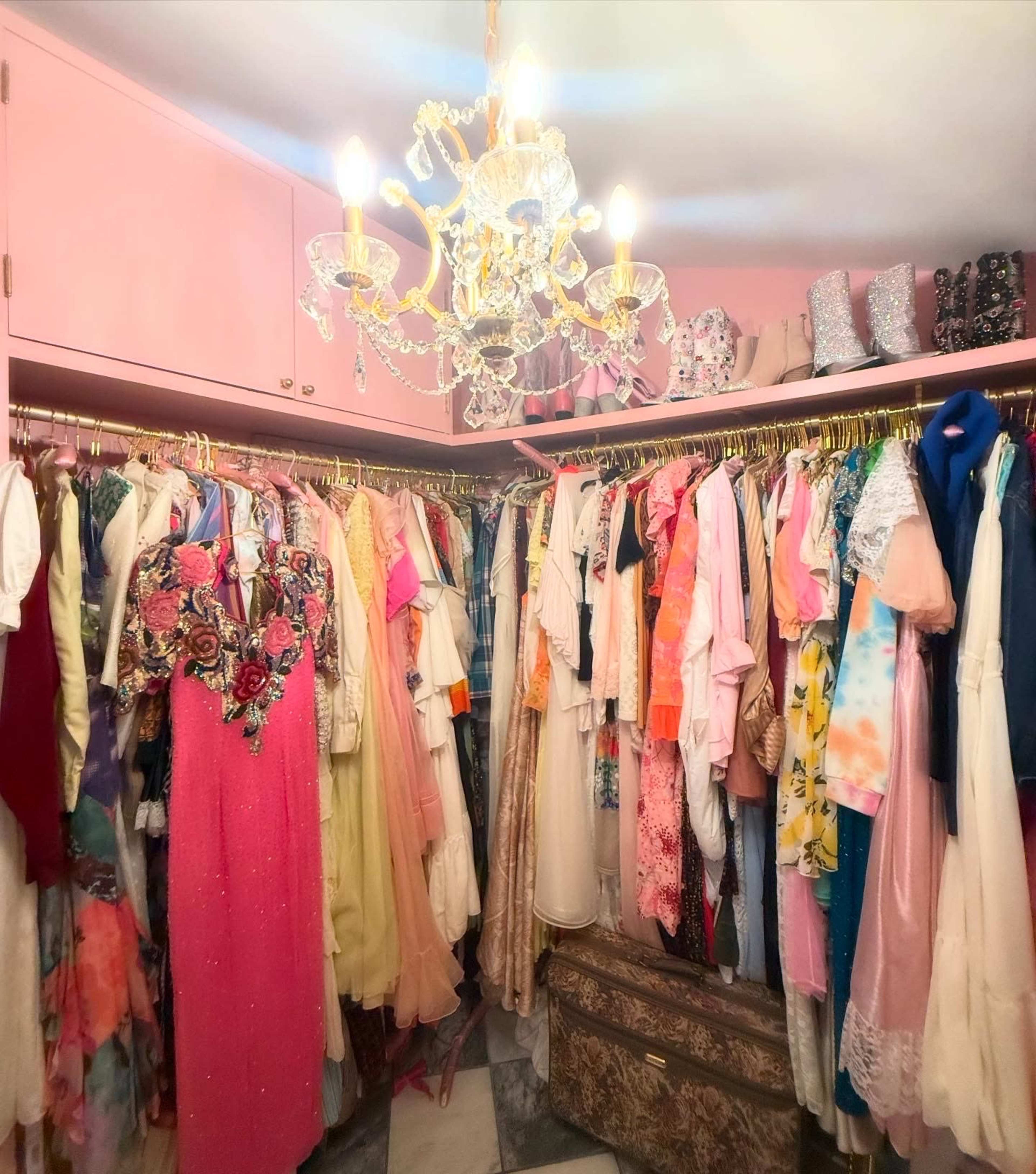 A brightly lit closet featuring a variety of colorful dresses and garments hanging on golden hangers, with a chandelier above.