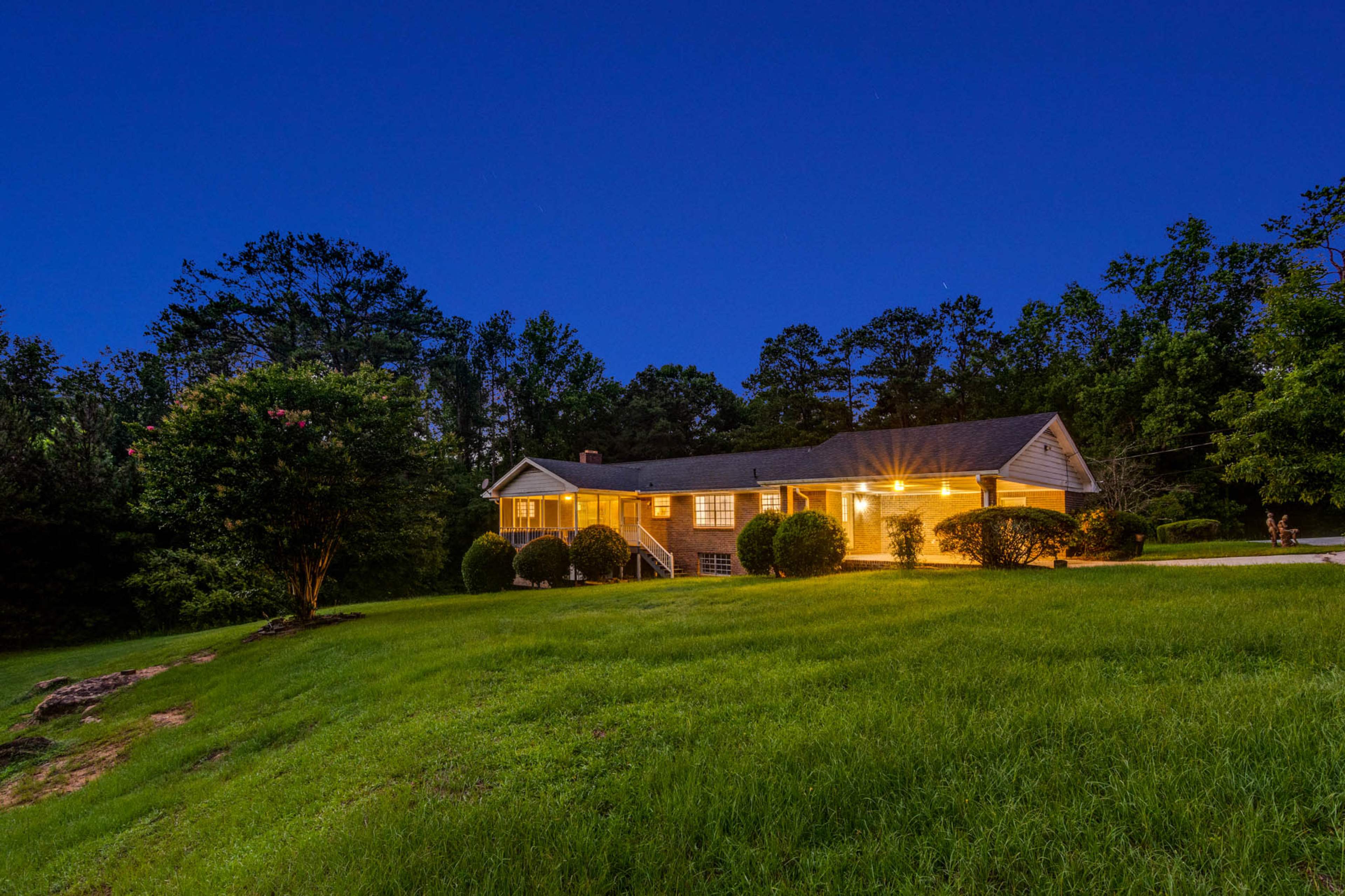A single-story house is illuminated at dusk, surrounded by grassy land and trees.