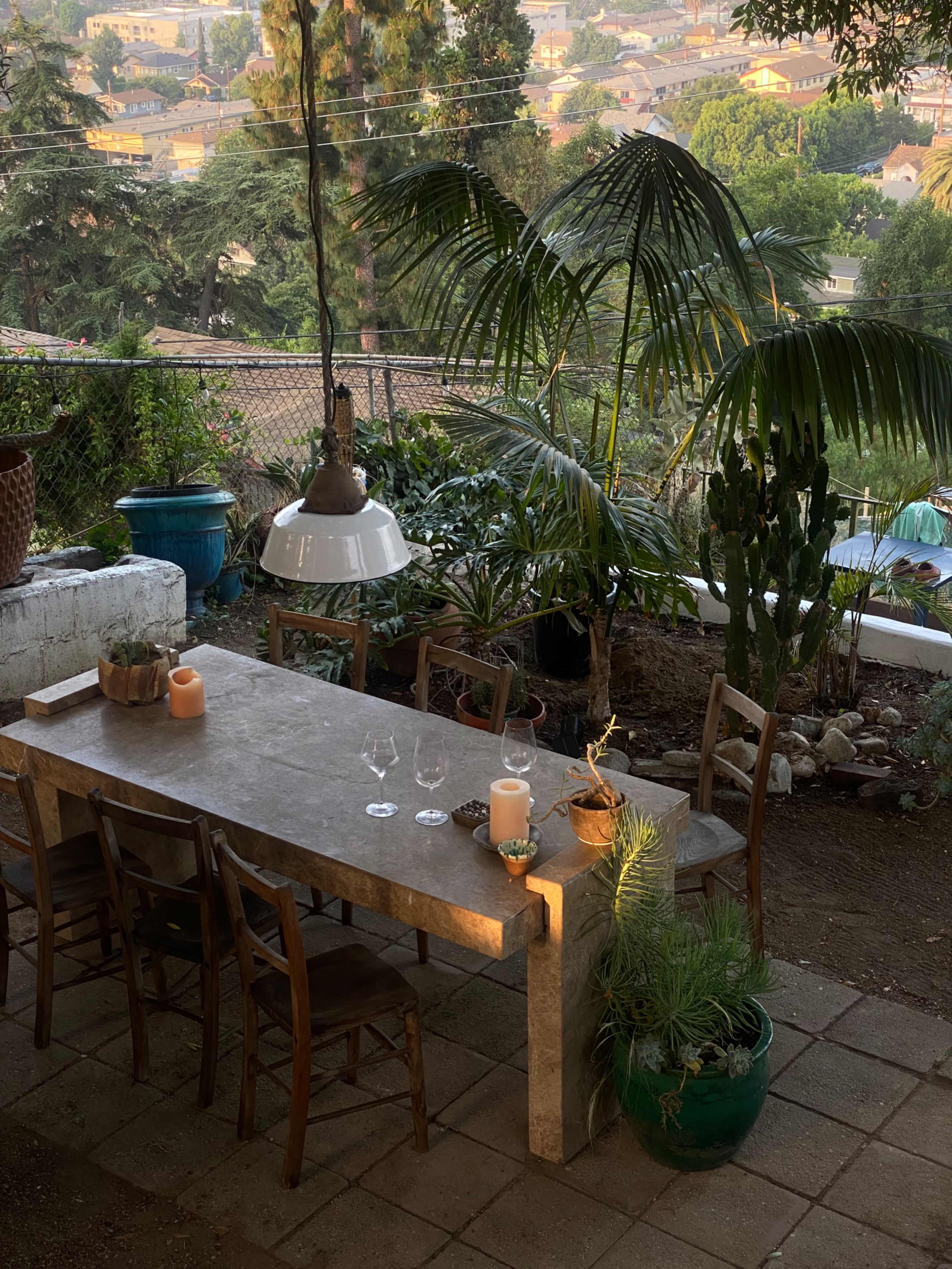A stone dining table with wooden chairs is set outdoors among various plants and trees, illuminated by a hanging light fixture.