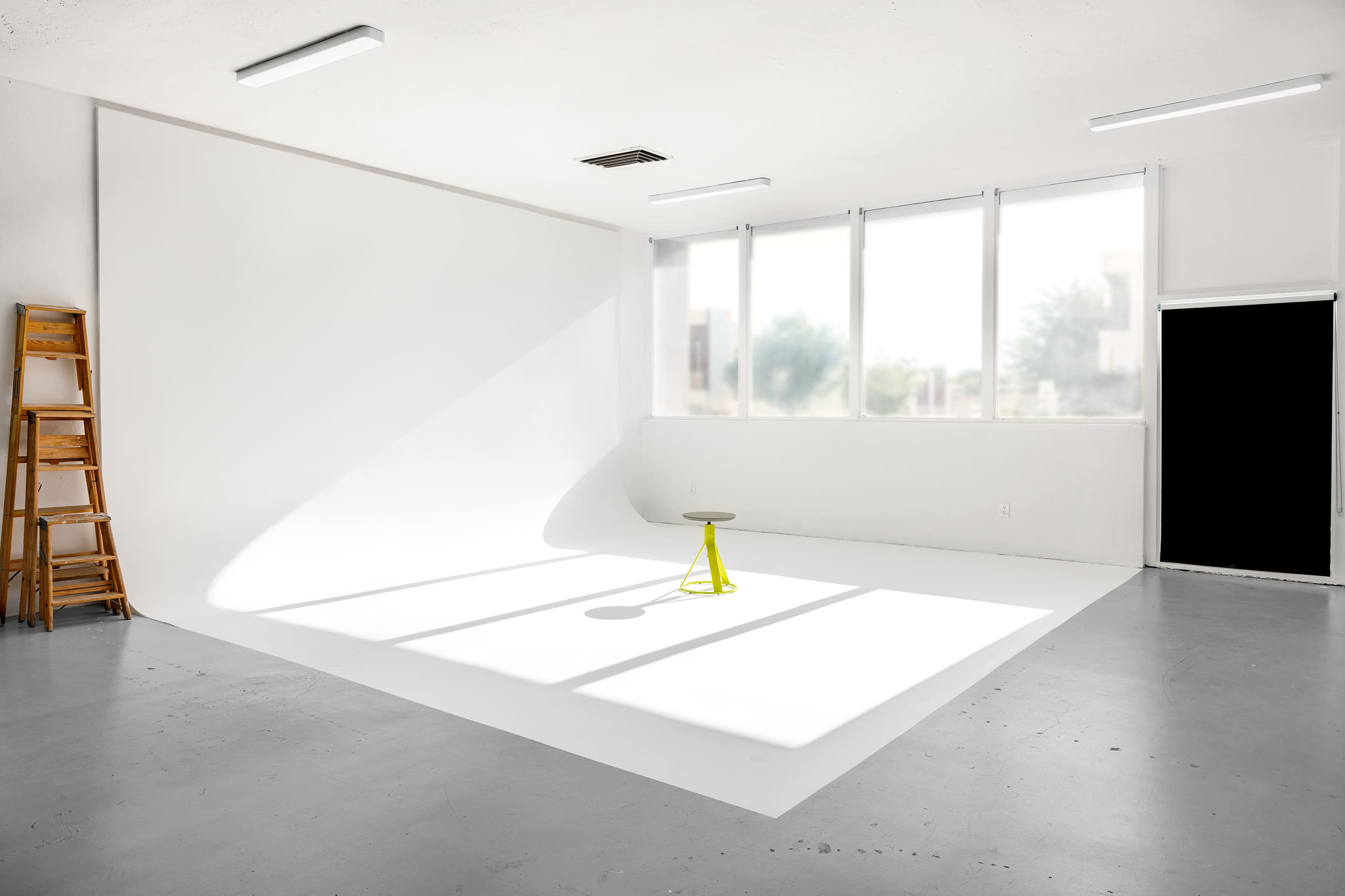 A minimalist photography studio features a yellow stool on a white backdrop with natural light streaming through large windows.