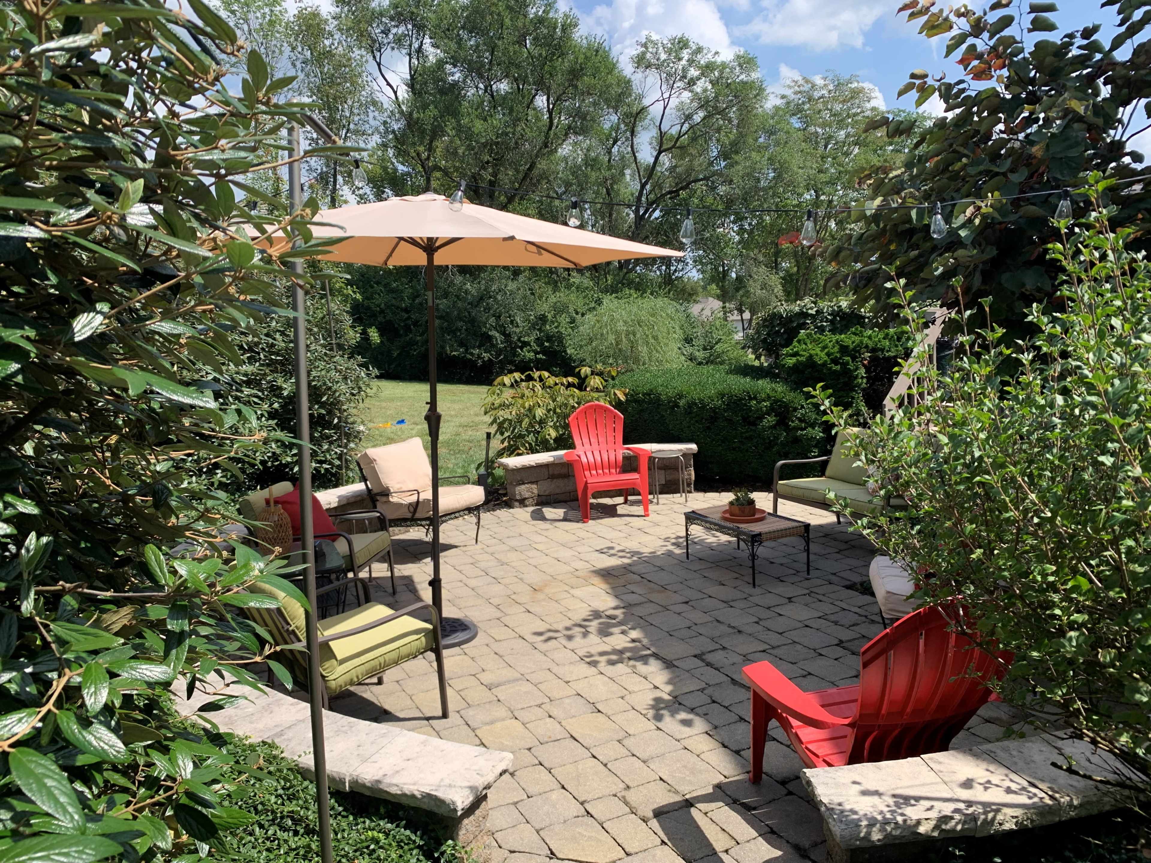 A landscaped patio with seating arrangements, an umbrella, and a stone fire pit surrounded by greenery.