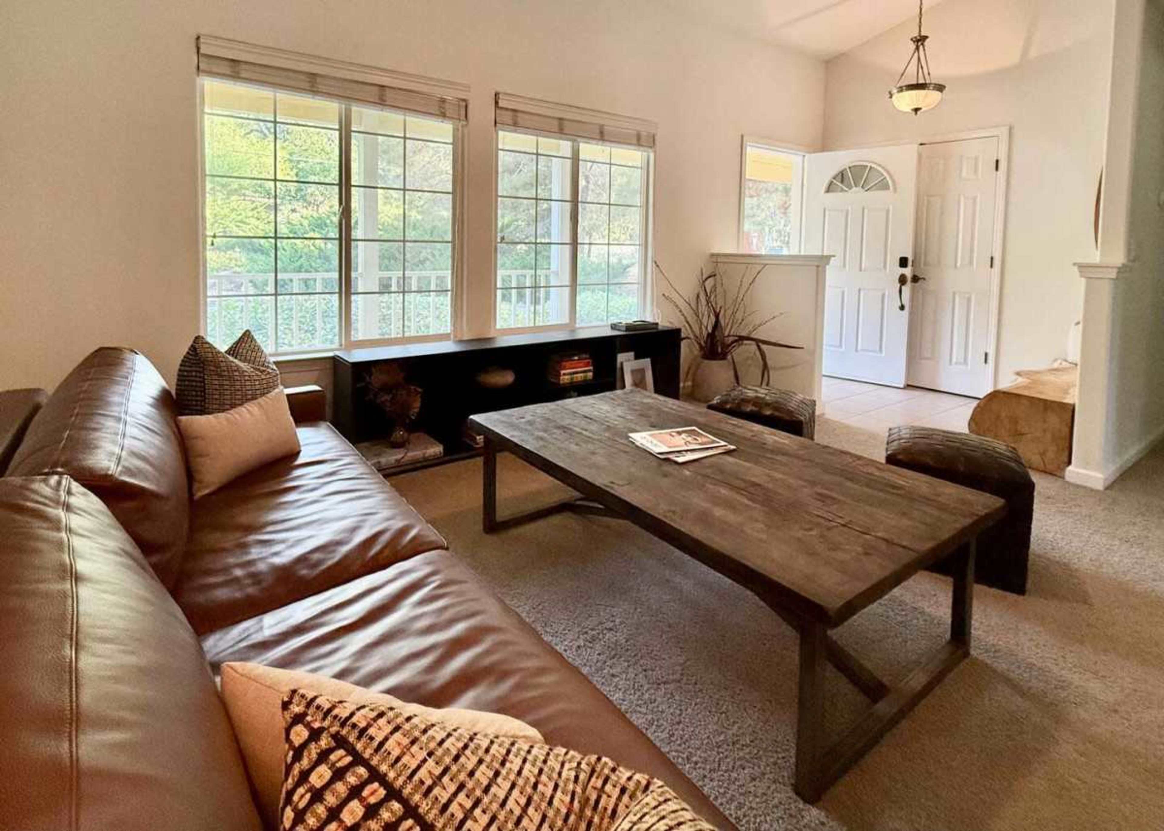 A living room features a brown leather sofa, a wooden coffee table, and large windows letting in natural light.