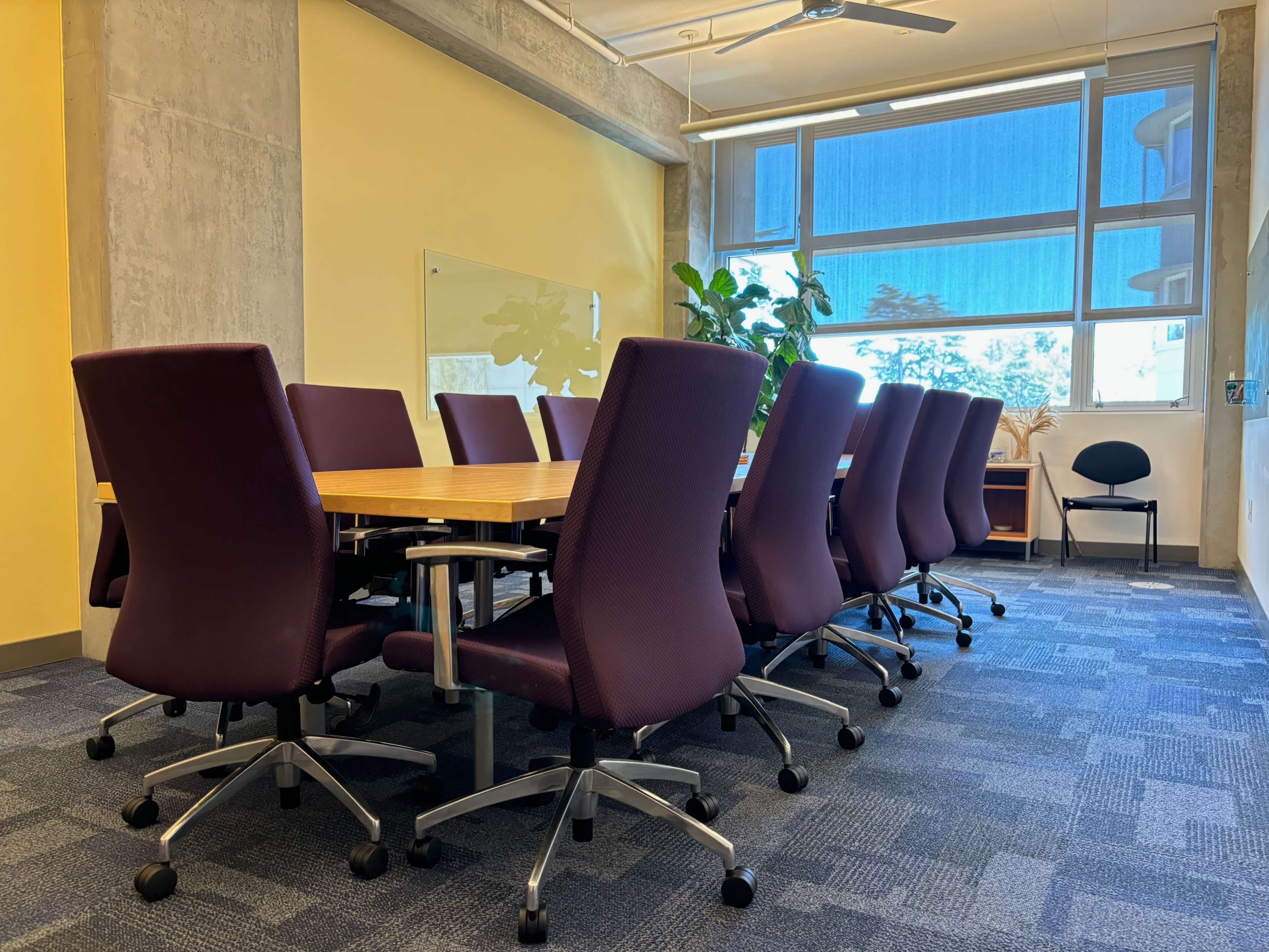 A conference room features a long wooden table surrounded by purple chairs and a large window letting in natural light.