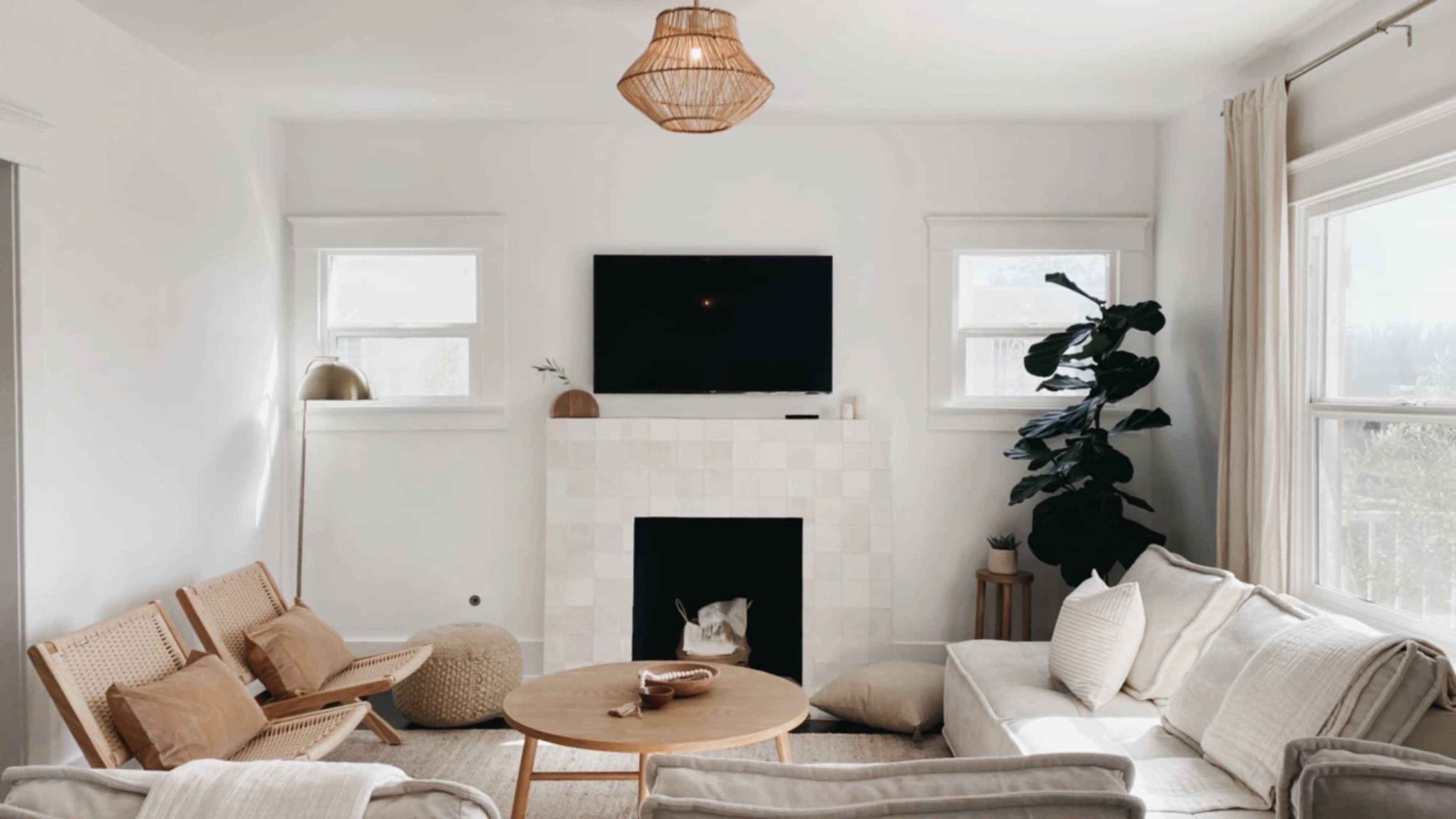 A cozy living room features a light-colored sofa and armchairs, a round wooden coffee table, and a fireplace with a TV above, surrounded by large windows and a potted plant.