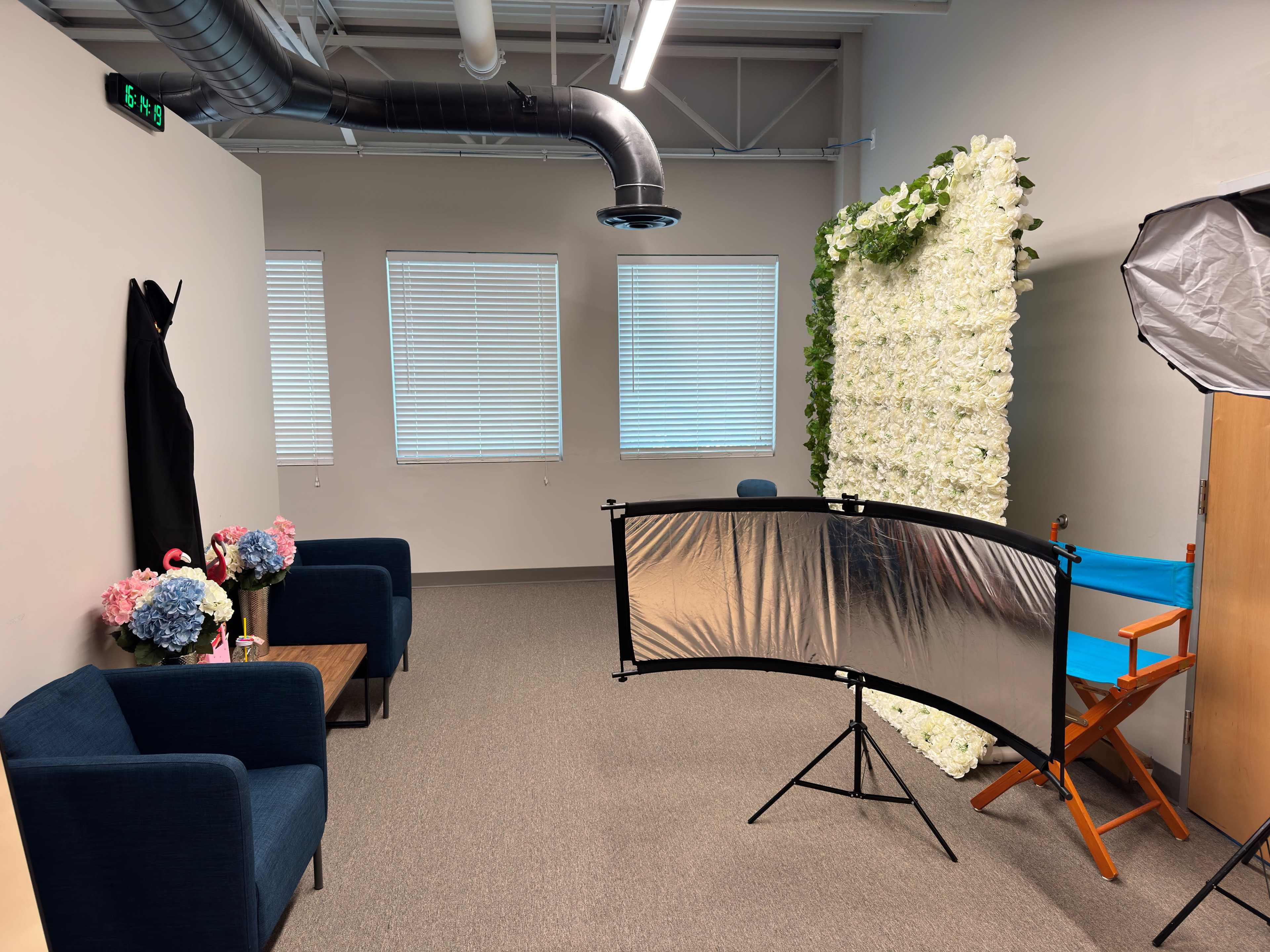 The image shows a well-lit room featuring two blue chairs, a small table with floral arrangements, a backdrop of artificial flowers, and a reflector setup for photography.