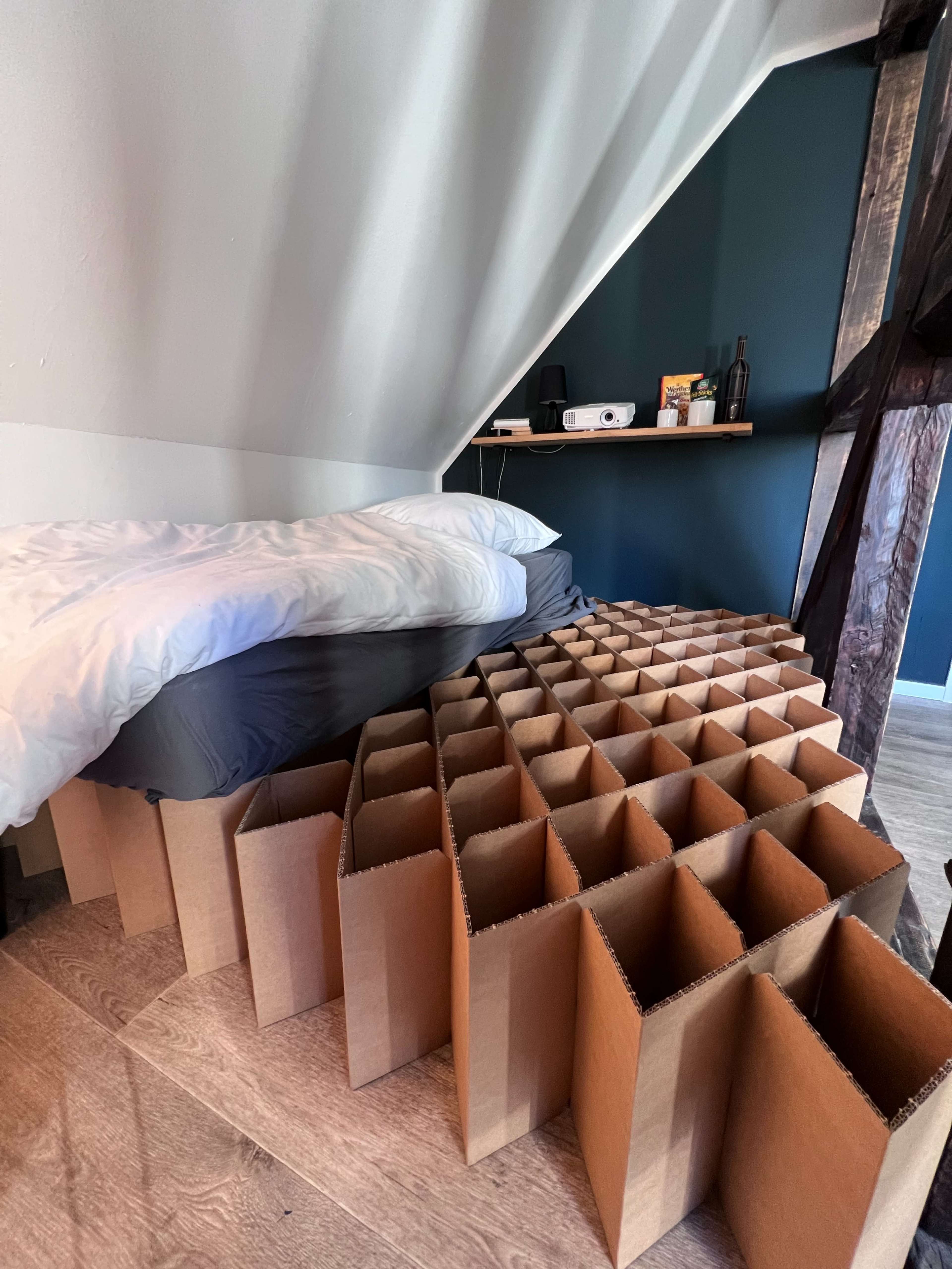 The image shows a bed with white and gray bedding positioned next to a structured arrangement of cardboard boxes on a wooden floor.