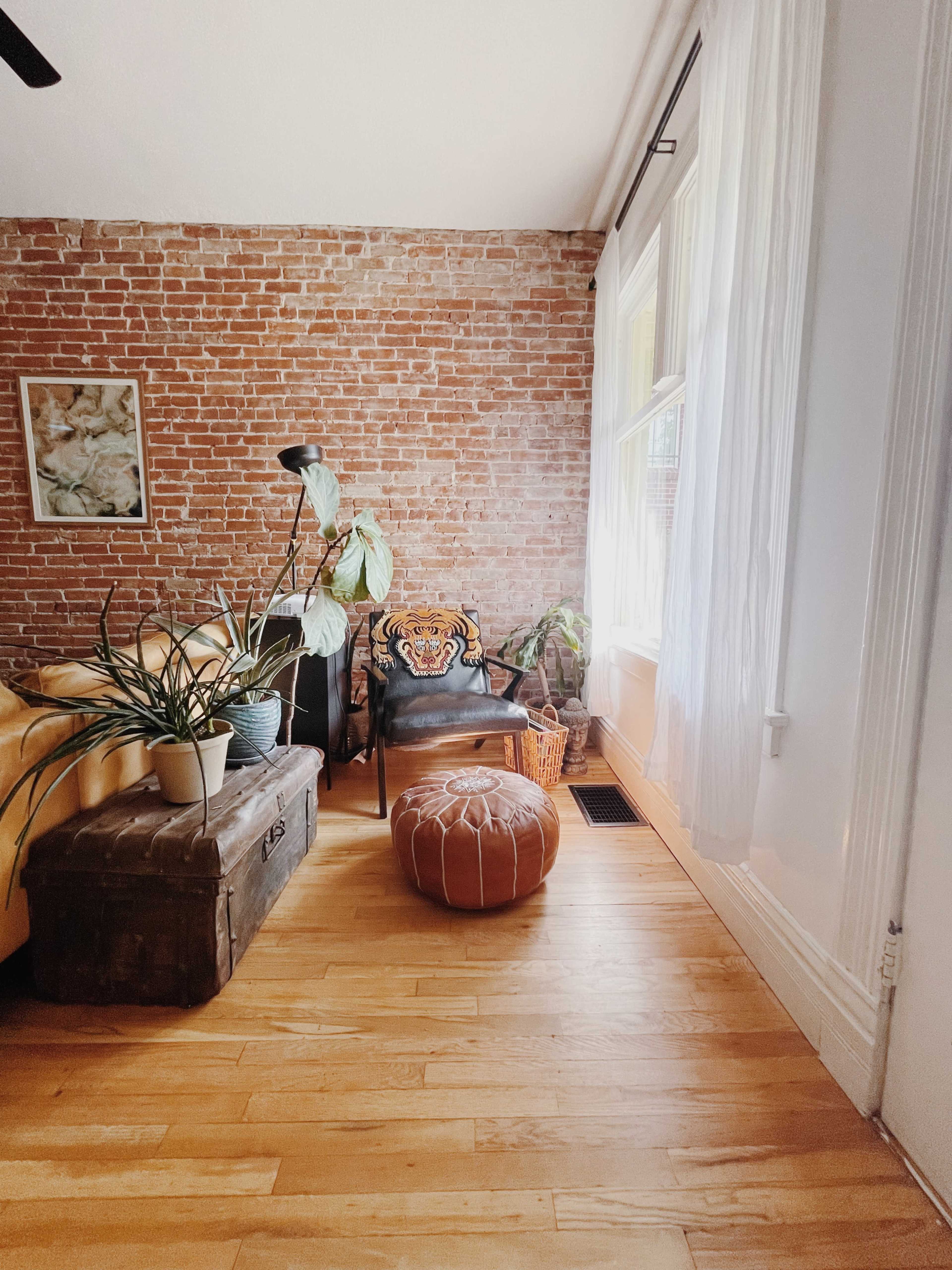 A cozy living room with brick walls, a yellow sofa, a wooden trunk, a plant, and a patterned pouf.