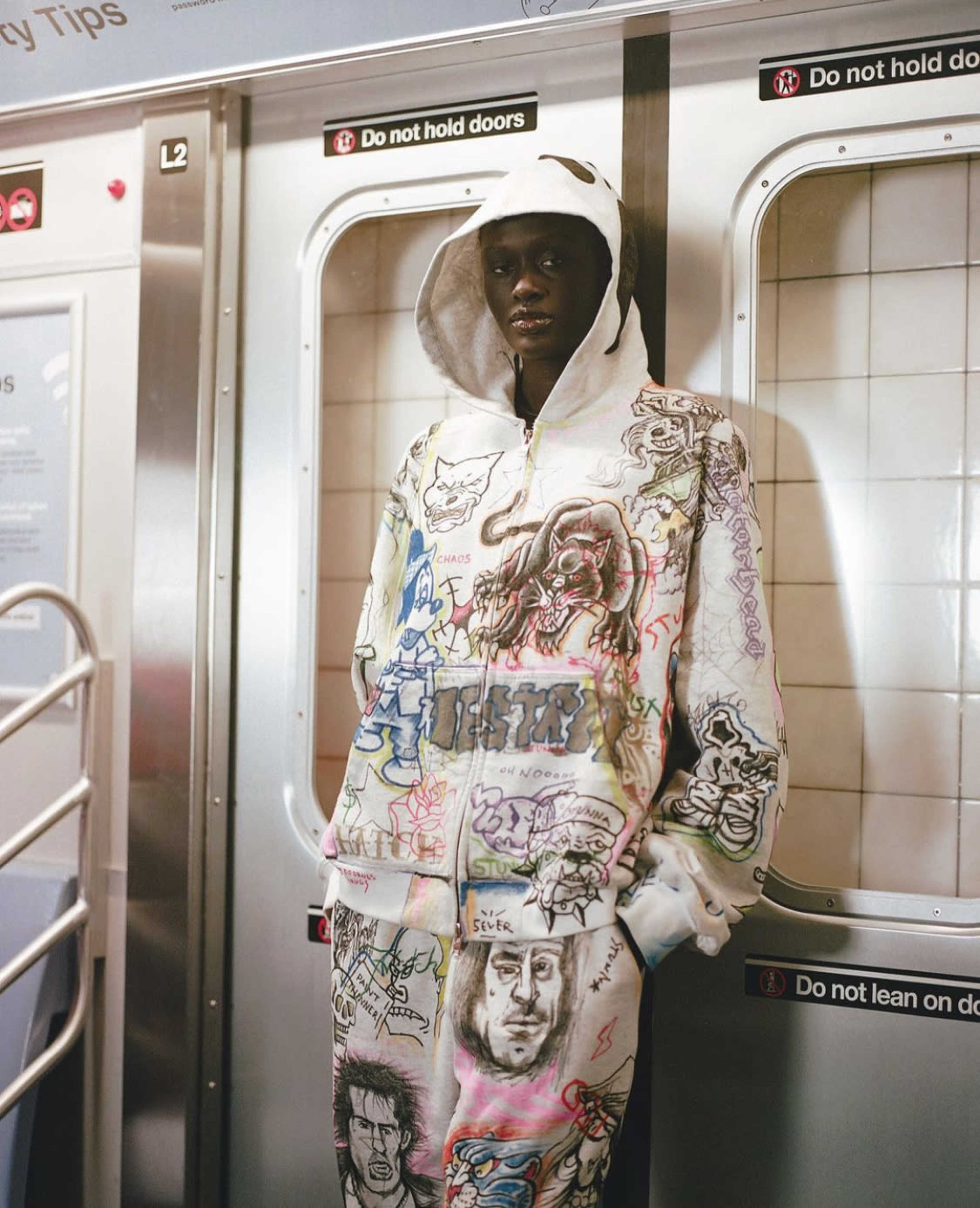 A person wearing a graffiti-style hoodie and sweatpants stands inside a subway train near the doors.