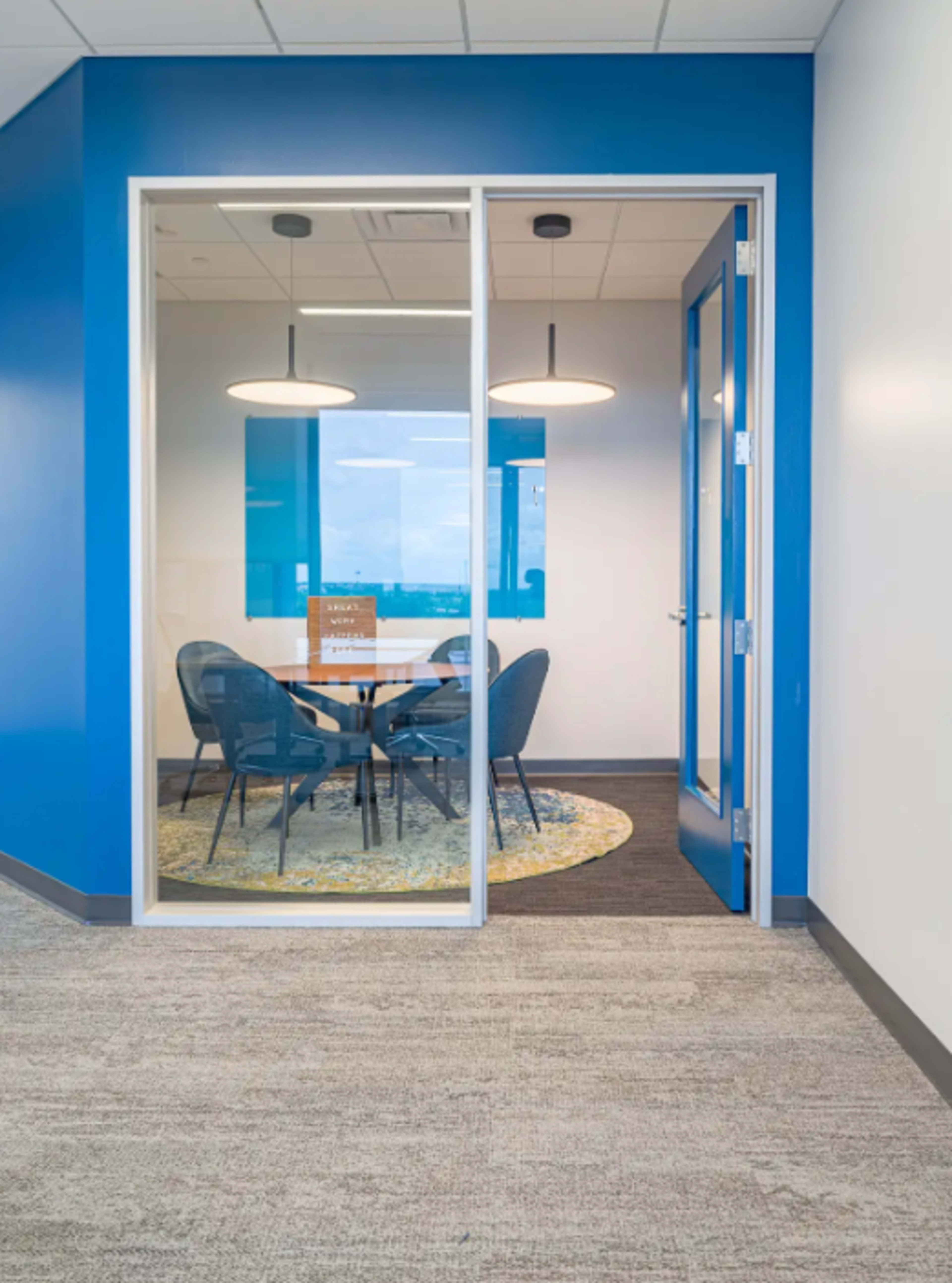 A modern conference room with a round table and four chairs is visible through a glass wall, accented by blue walls and overhead lighting.