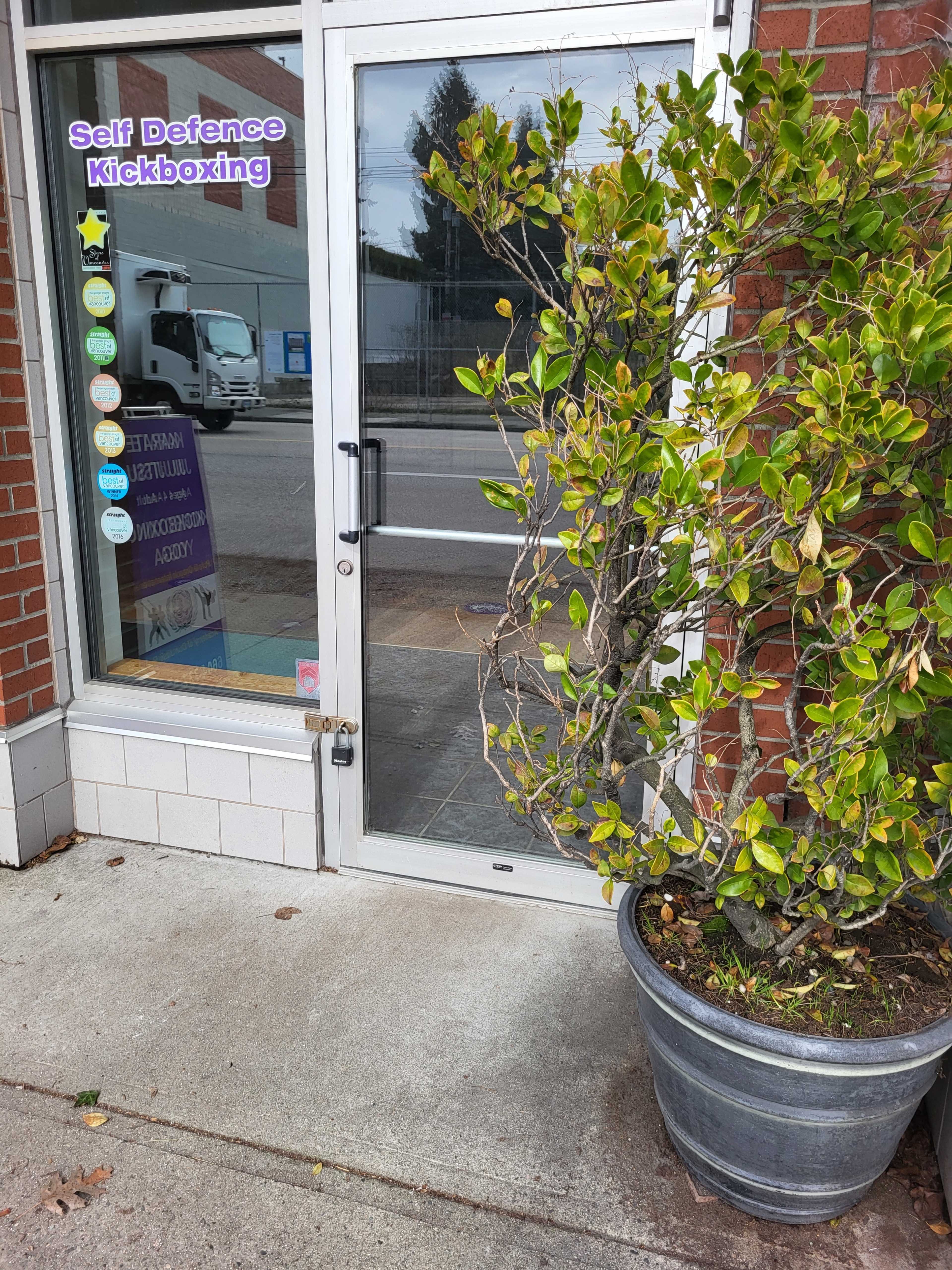 The image shows a glass door entrance to a self-defense kickboxing studio, flanked by a large potted plant on a concrete sidewalk.