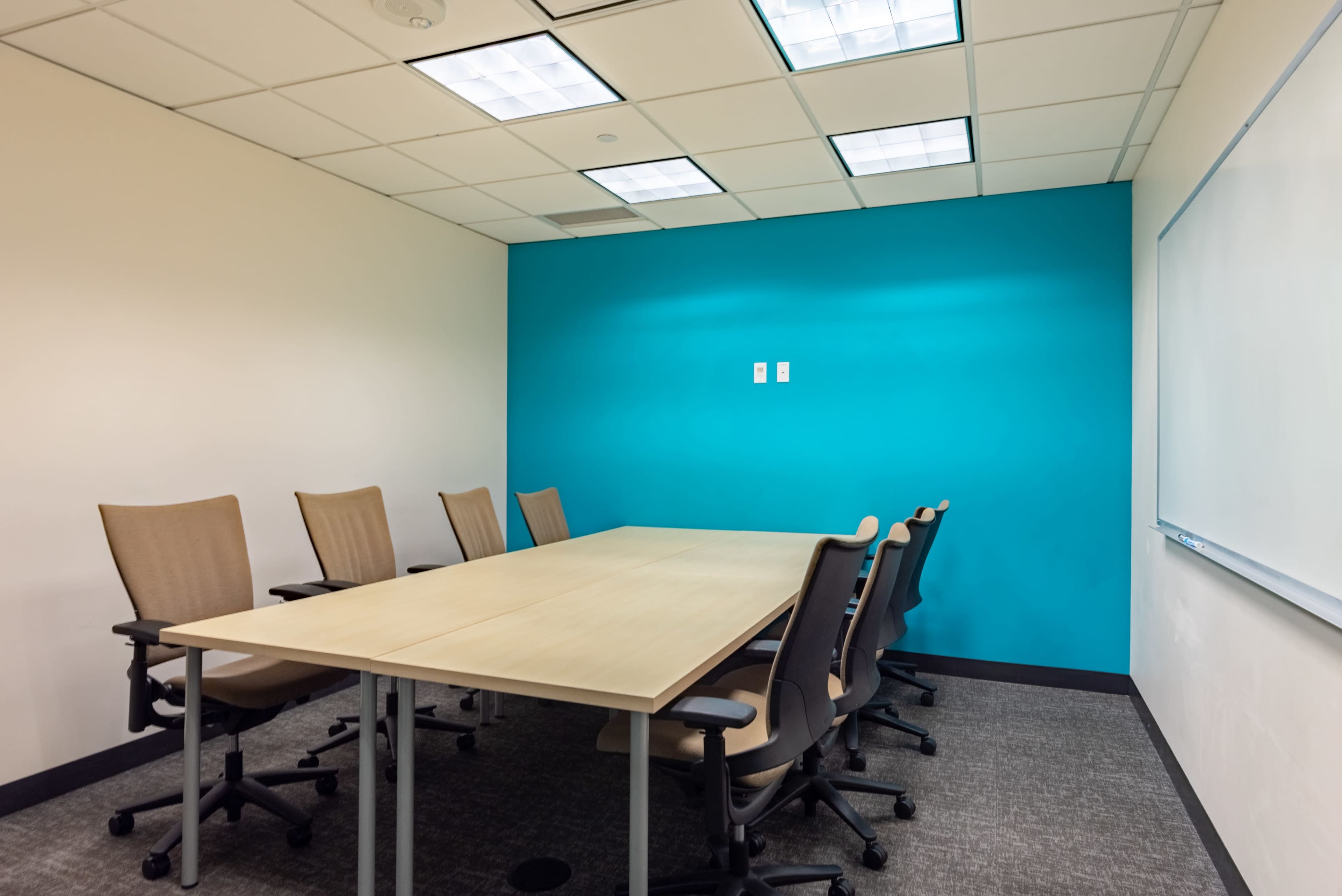 A conference room features a wooden table surrounded by eight beige chairs, with a teal accent wall and a whiteboard.