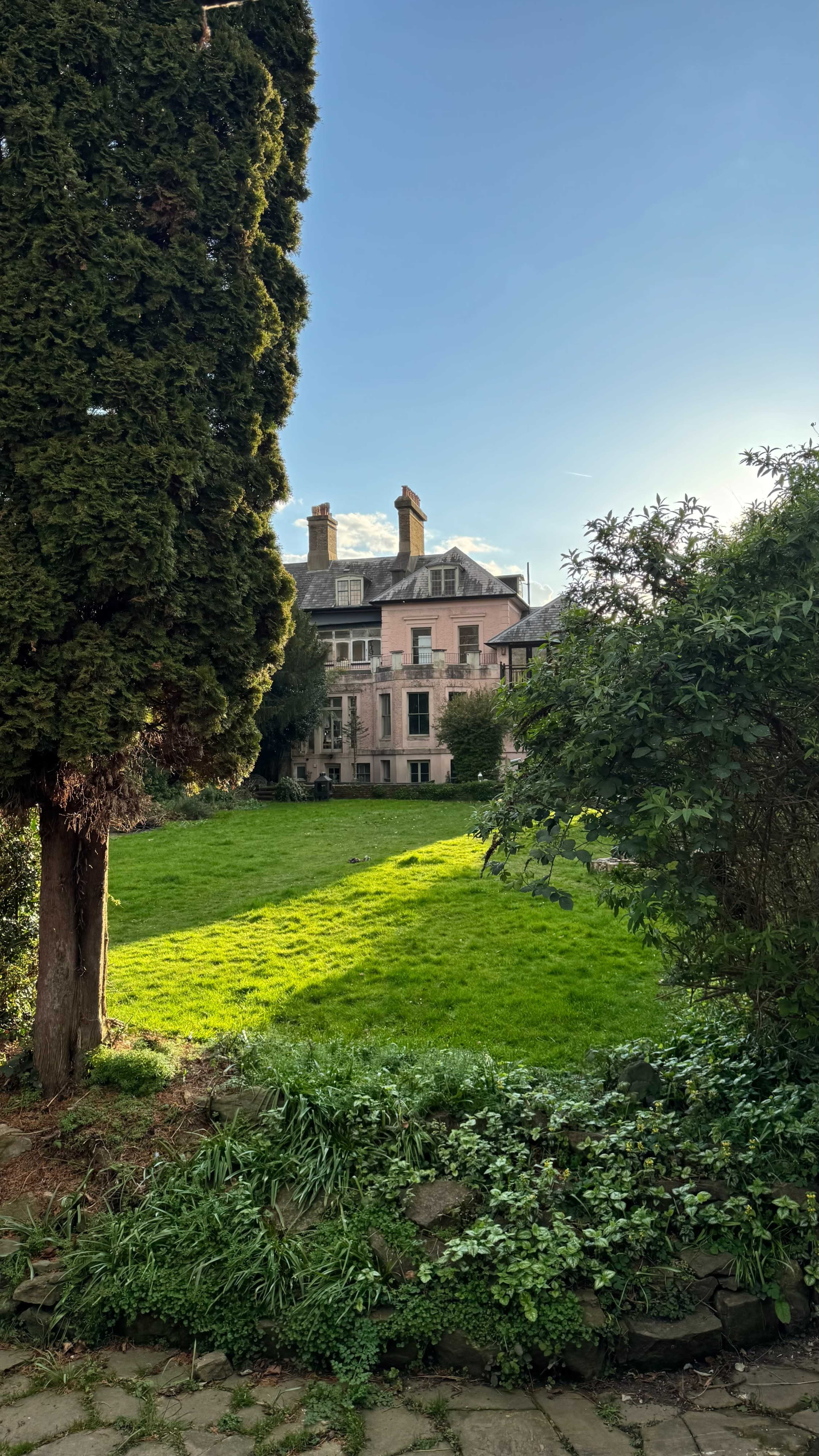 A large, pink mansion is set against a clear sky, surrounded by lush green grass and trees.
