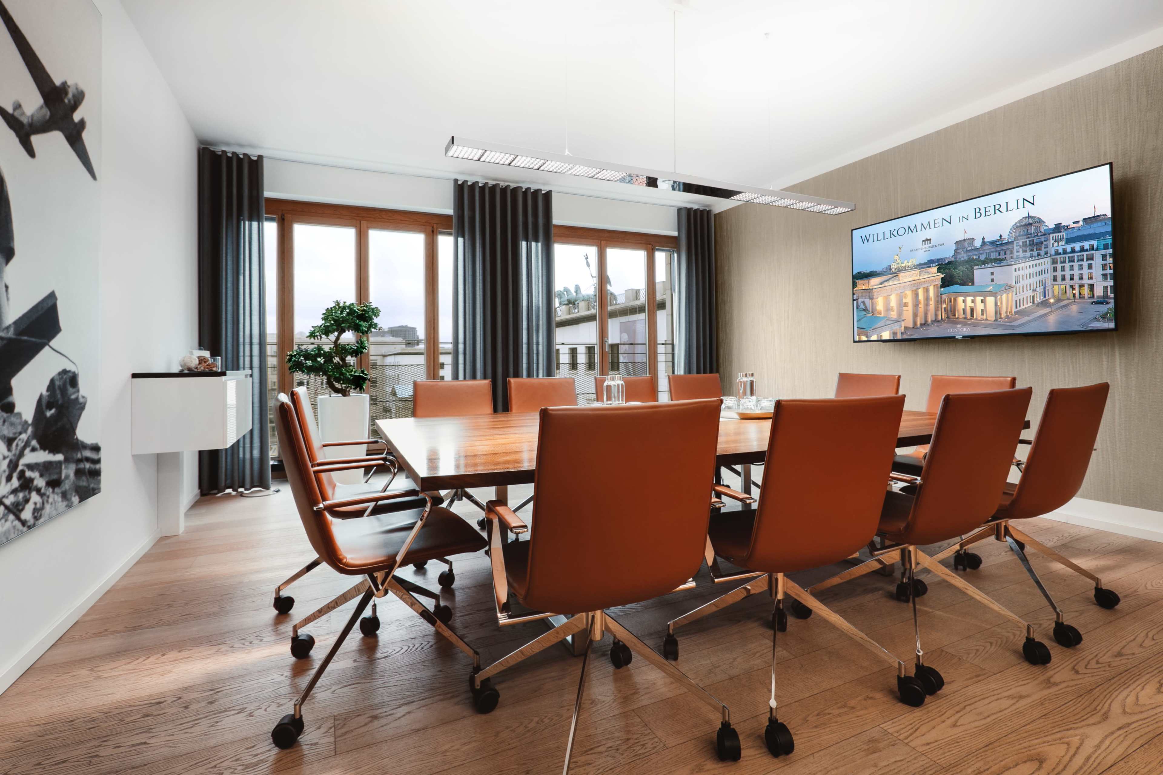 A spacious conference room features a large wooden table surrounded by orange leather chairs, with a large screen displaying a welcome message in Berlin and windows providing natural light.