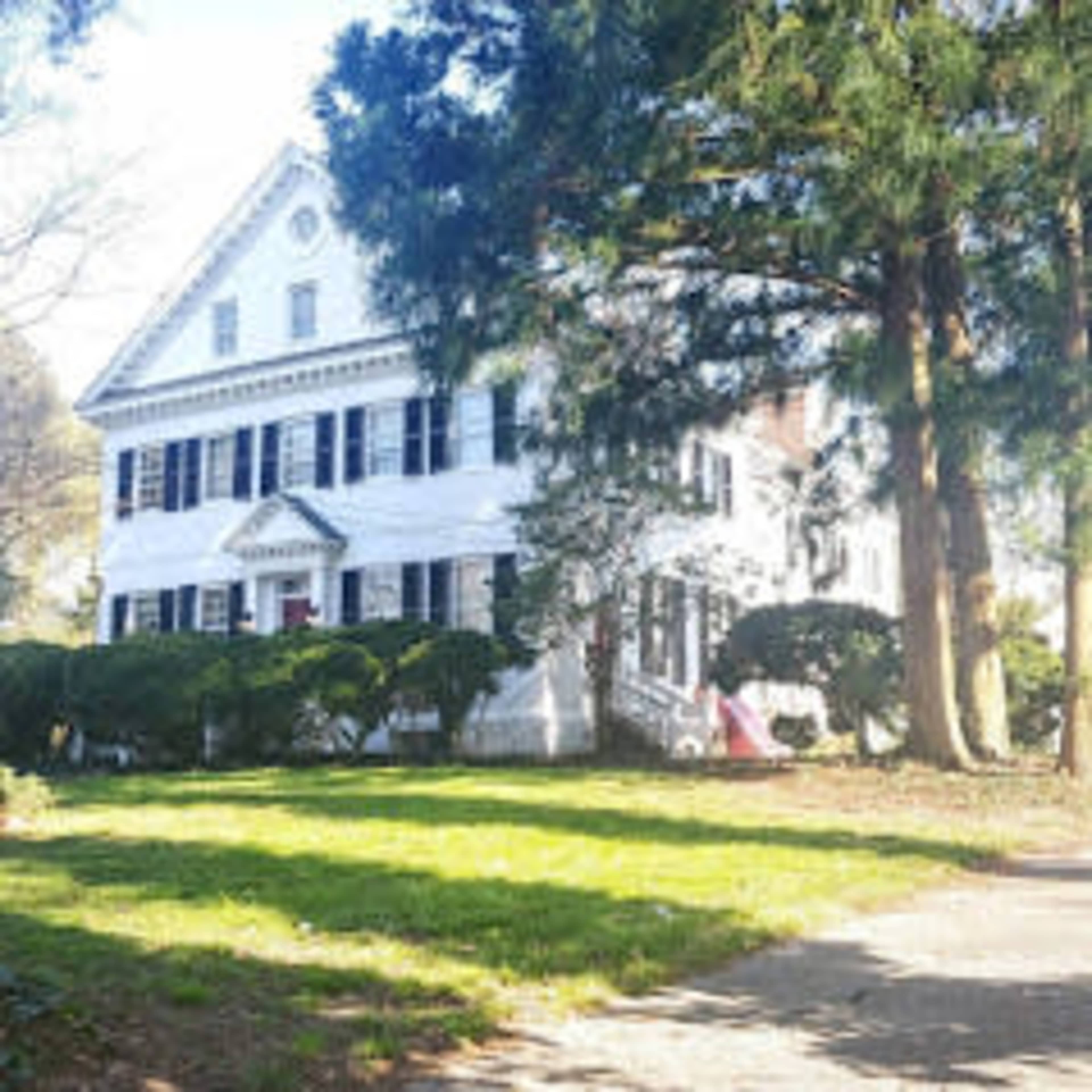 A large white house with black shutters surrounded by greenery and trees.