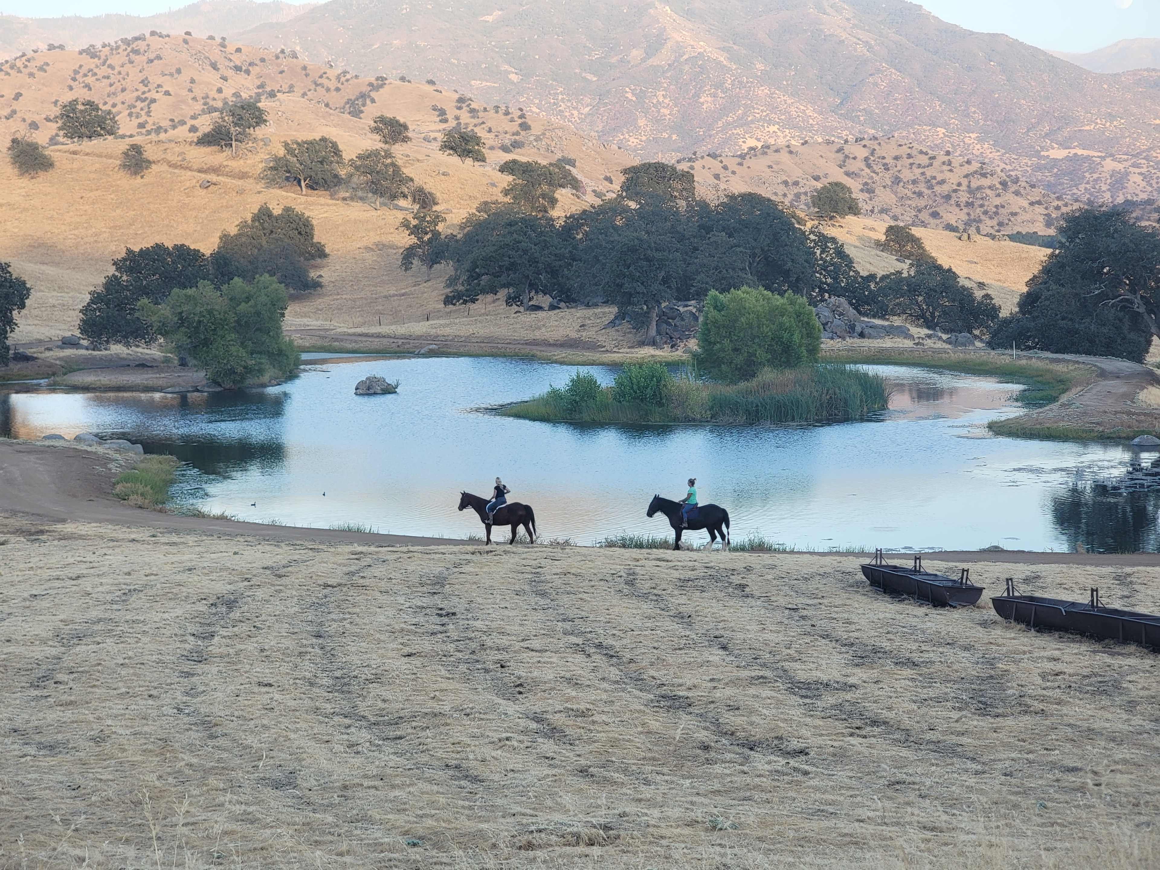 Two riders on horseback are near a pond surrounded by rolling hills and sparse trees.