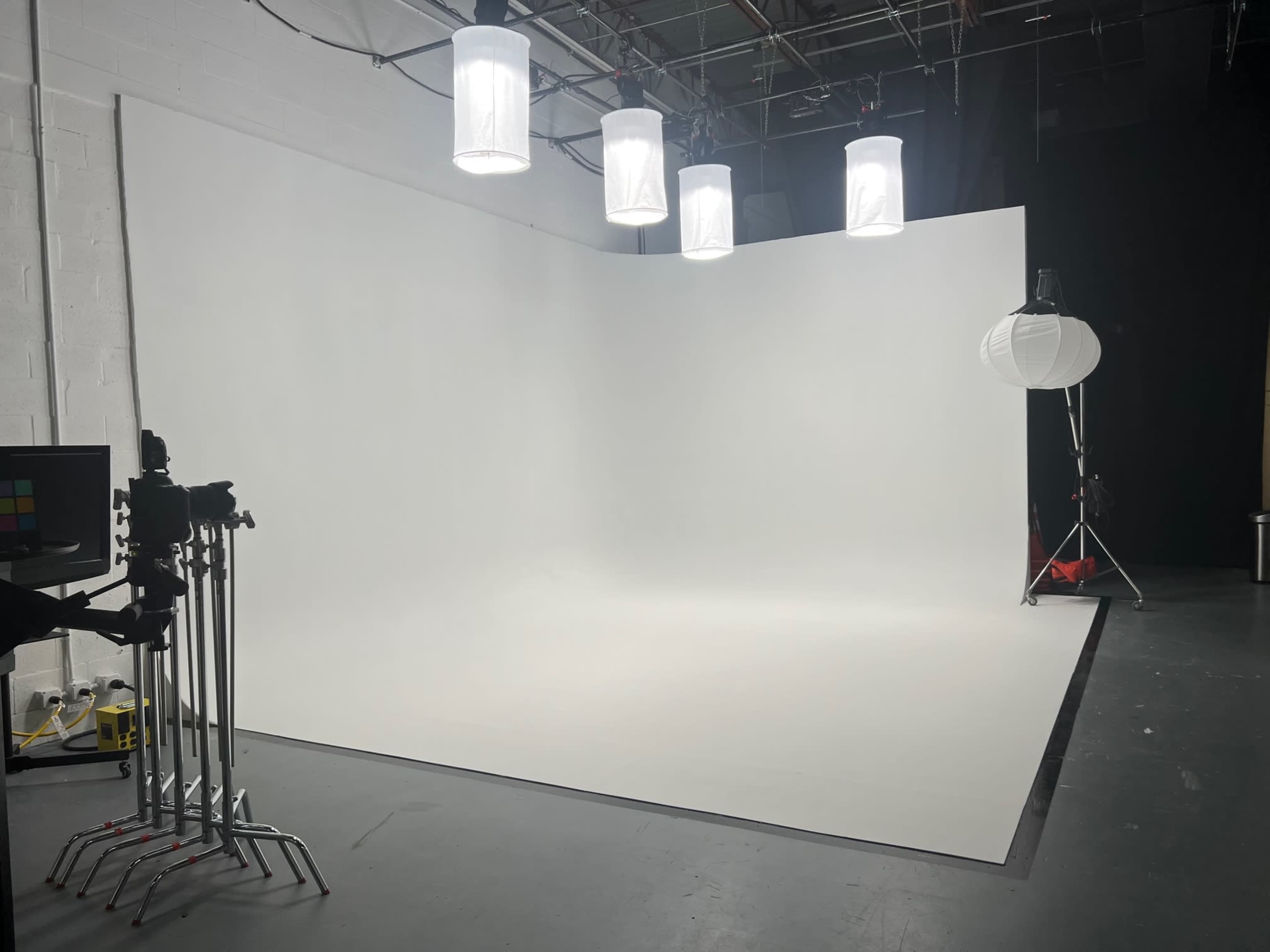 A photography studio featuring a seamless white backdrop, multiple hanging lights, and camera equipment on tripods.