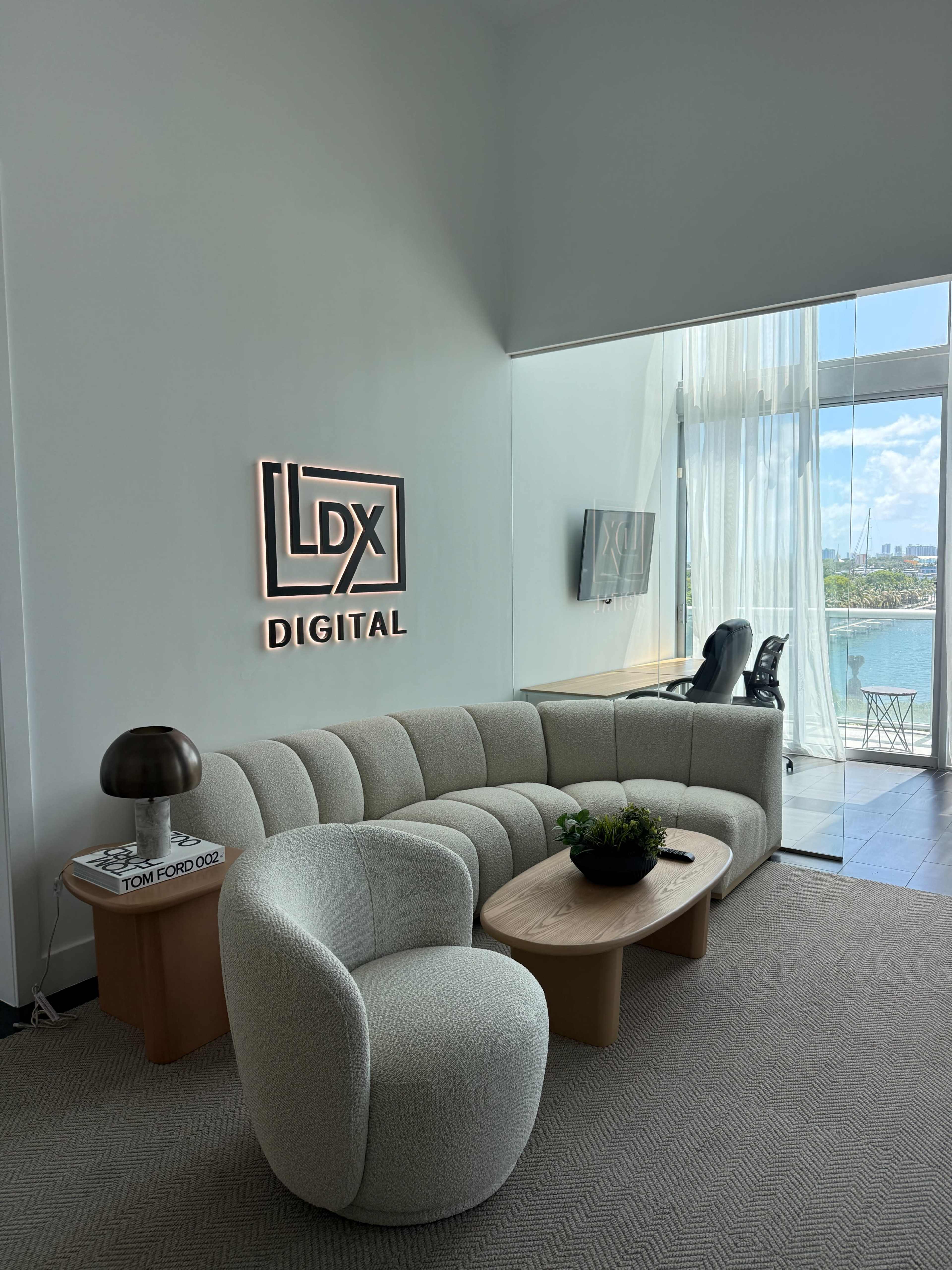 The image shows a modern lounge area featuring a curved gray sofa, a round coffee table with a plant, and a large window offering a view of the outside.
