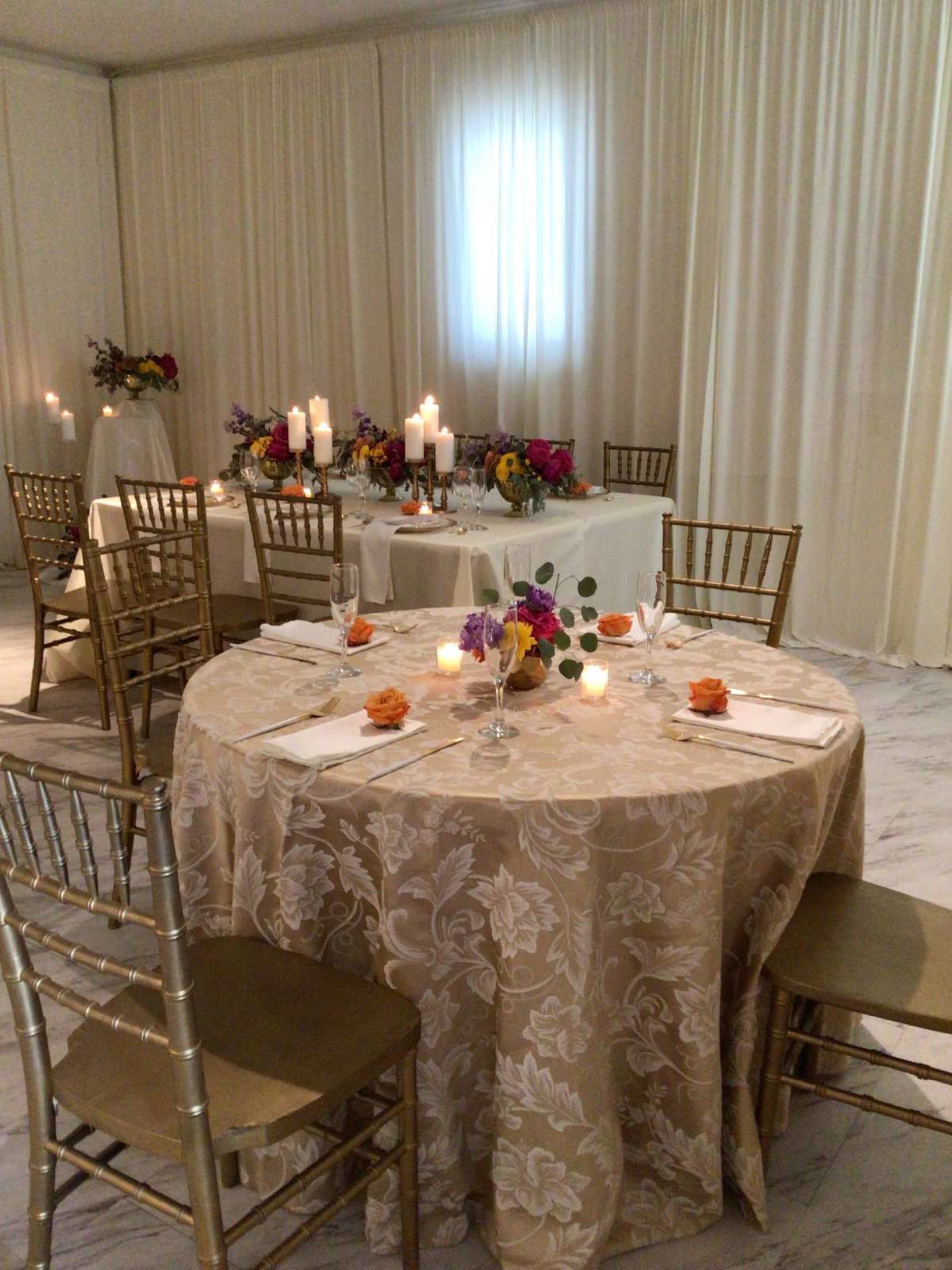 The image shows a formal dining setup with a round table covered in a floral tablecloth, surrounded by gold chairs and decorated with candles and floral centerpieces.