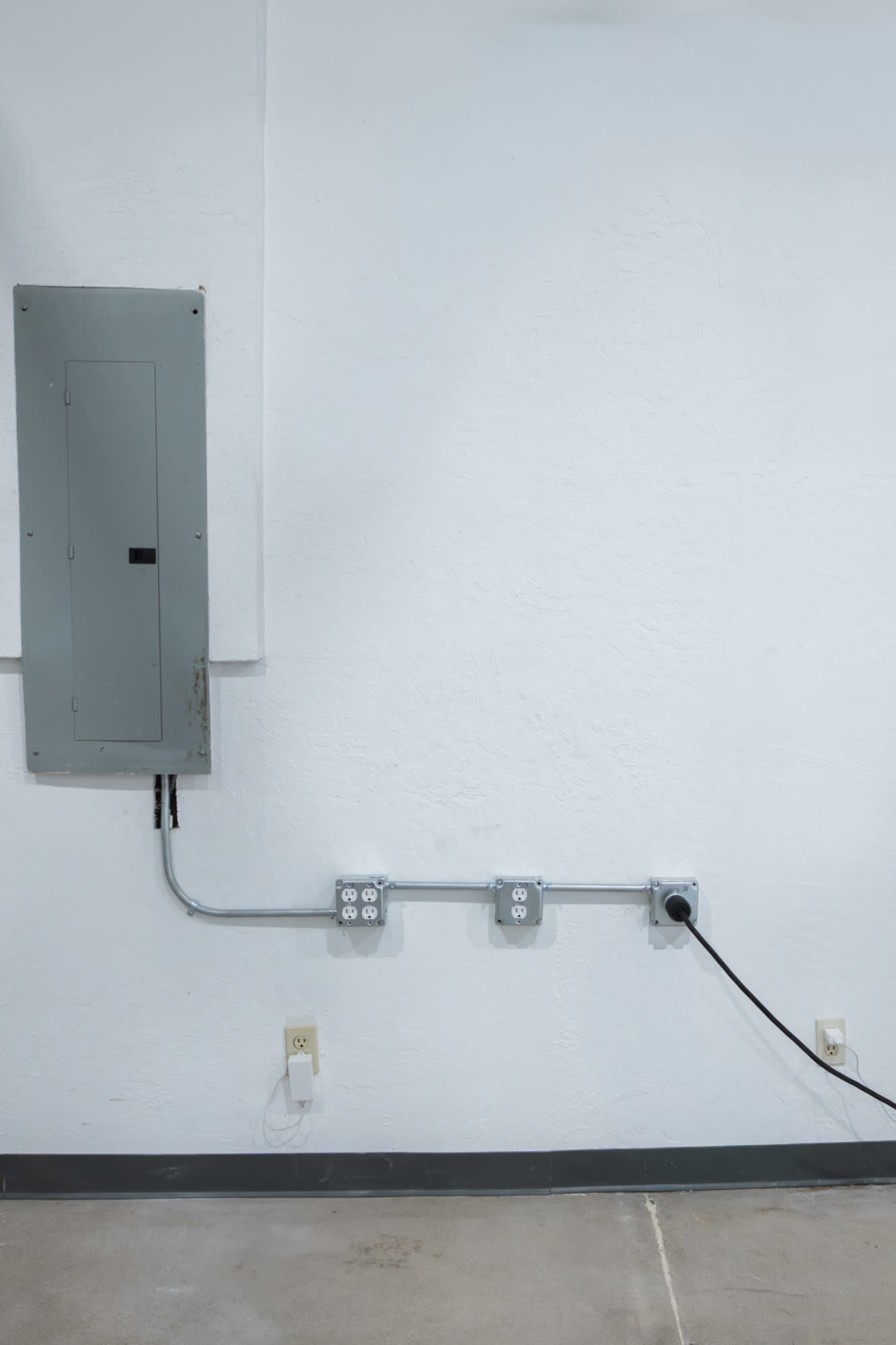 The image shows a gray electrical panel mounted on a white wall with two metal conduit boxes and an electrical outlet below it.