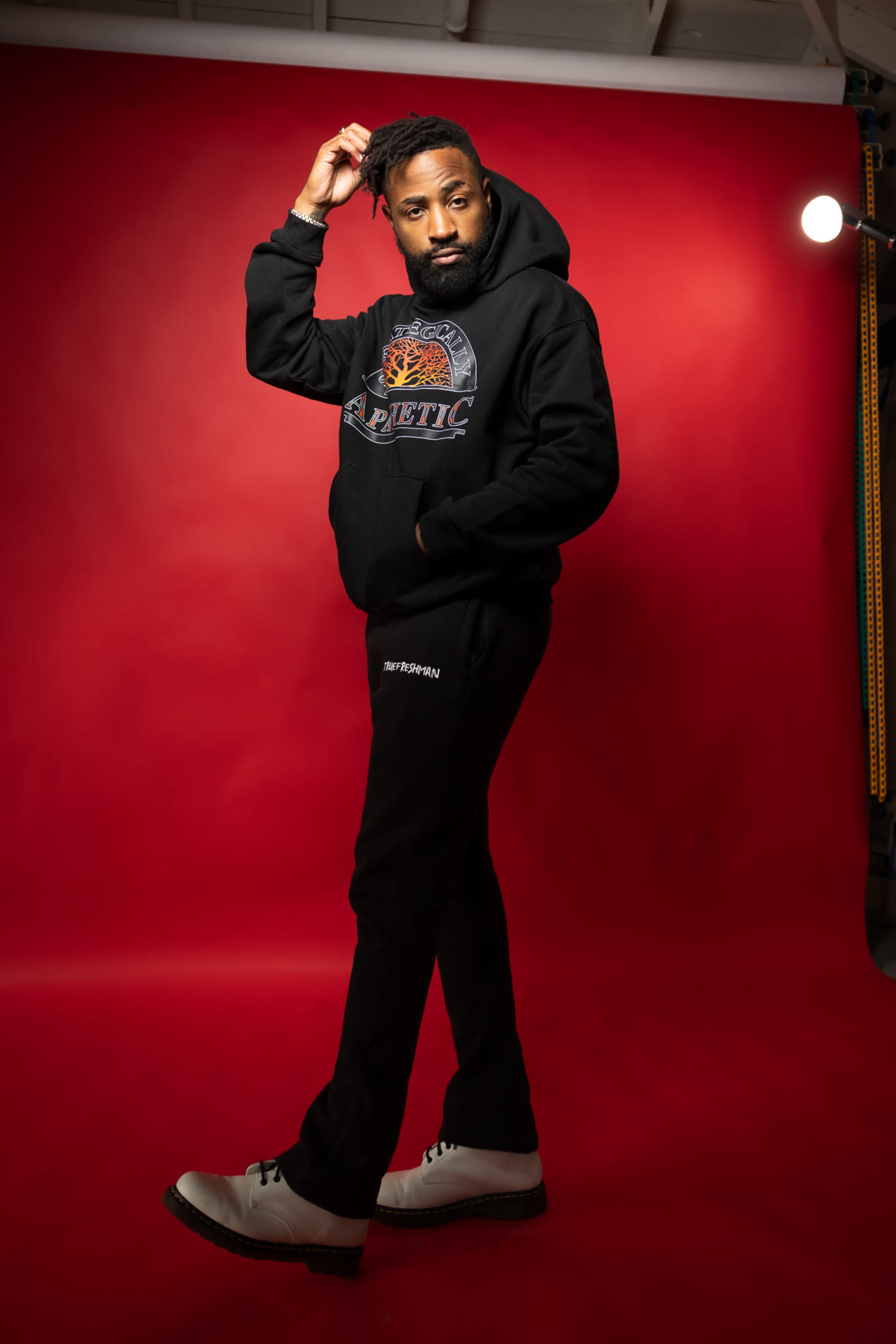 A man poses in front of a red backdrop, wearing a black hoodie and sweatpants with contrasting white shoes.
