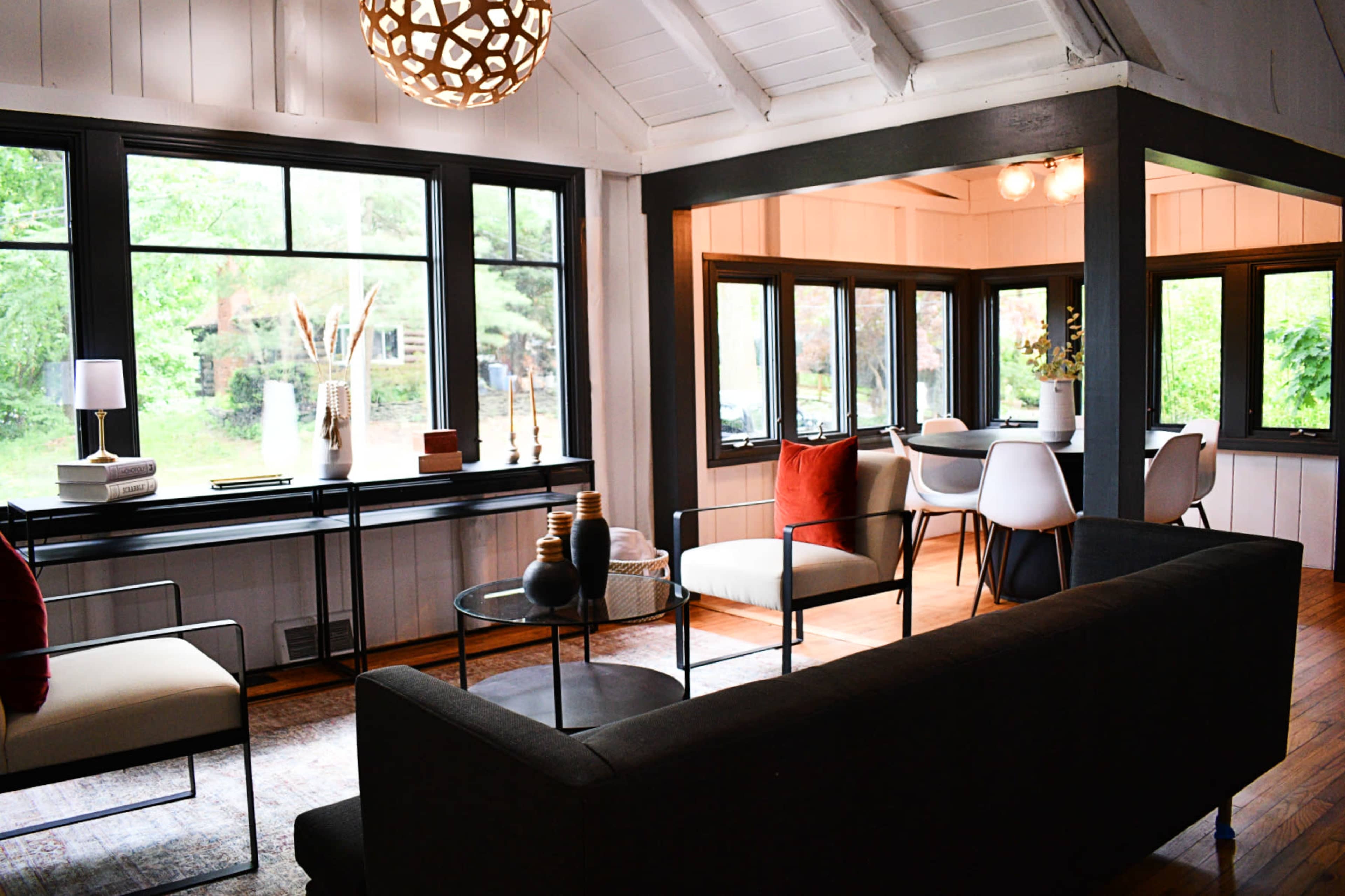 A modern living room with a dark sofa, two chairs, a coffee table, and large windows overlooking a green outdoor space.