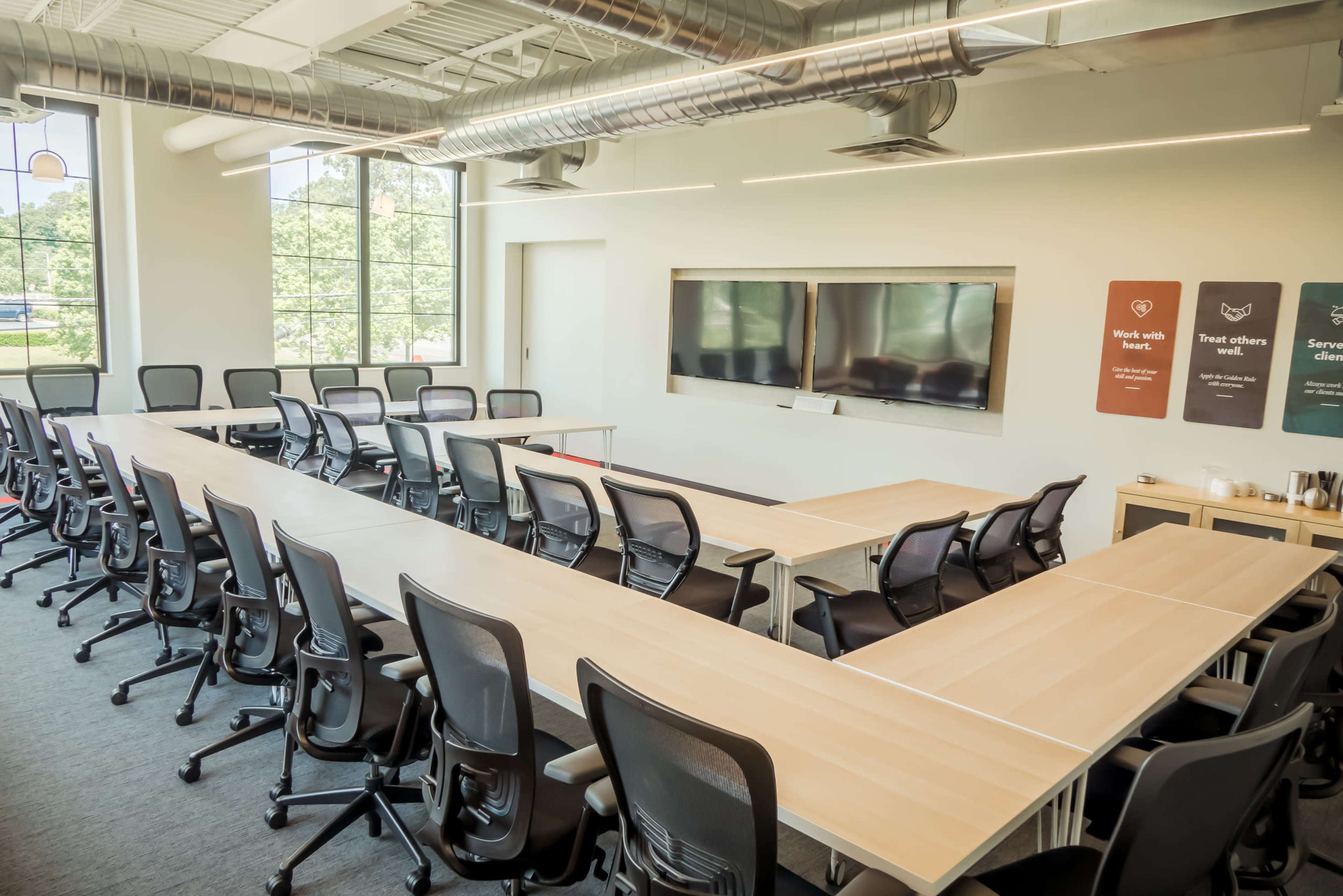 A modern conference room features a long U-shaped wooden table surrounded by ergonomic chairs, with large screens mounted on the wall and large windows letting in natural light.